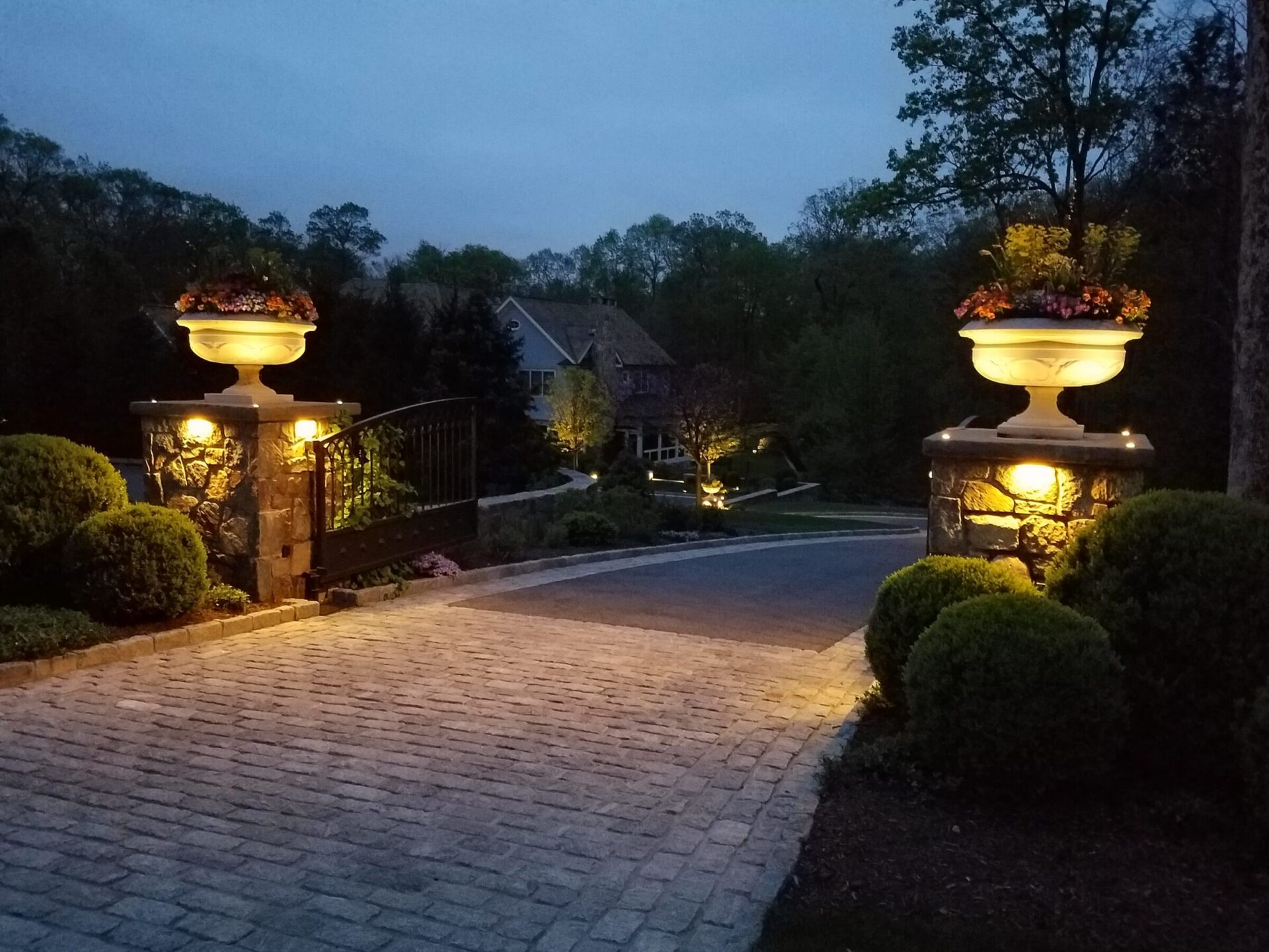 Elegant stone driveway entrance illuminated by warm landscape lighting, featuring large decorative urns with vibrant flowers atop stone pillars, leading to a home nestled in a wooded area at dusk.