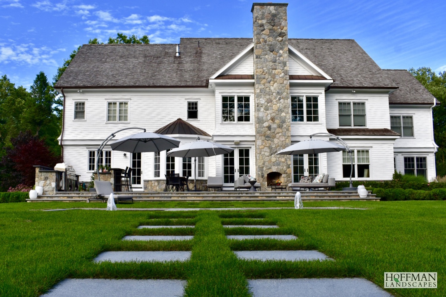 A large, elegant white house with a stone chimney, surrounded by manicured lawns, featuring outdoor seating areas with umbrellas, and a geometric garden path.