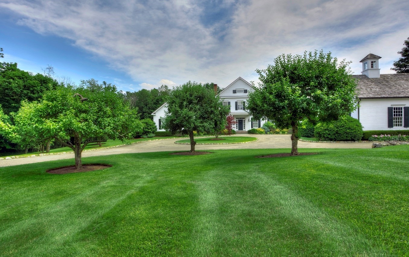 A large white house with a green lawn, trees, and a curved driveway under a partly cloudy blue sky. The property appears well-maintained and tranquil.