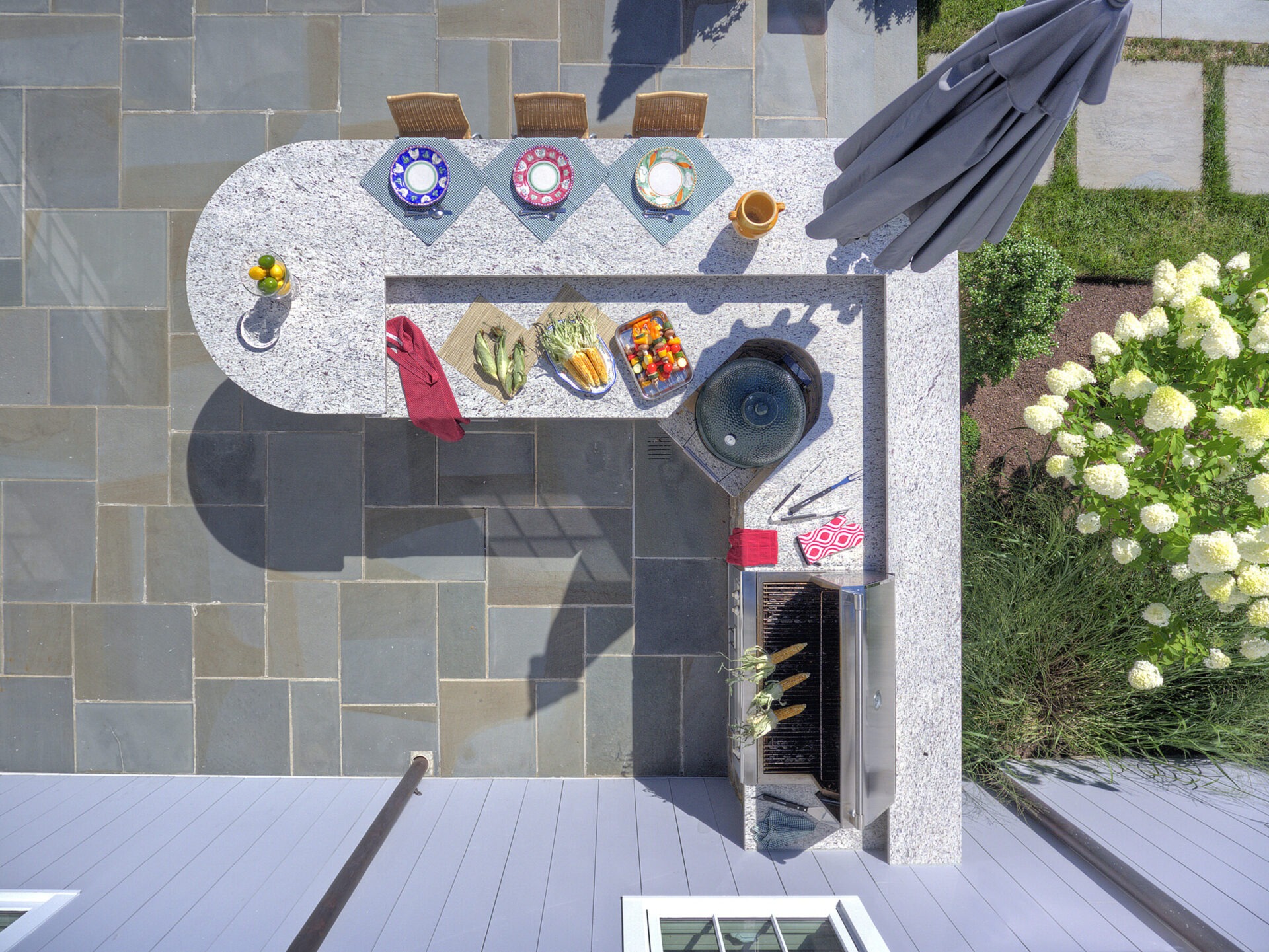 Aerial view of an outdoor kitchen counter with bar stools, a grill, plates, food, cooking utensils, a closed umbrella, and flowering bushes nearby.