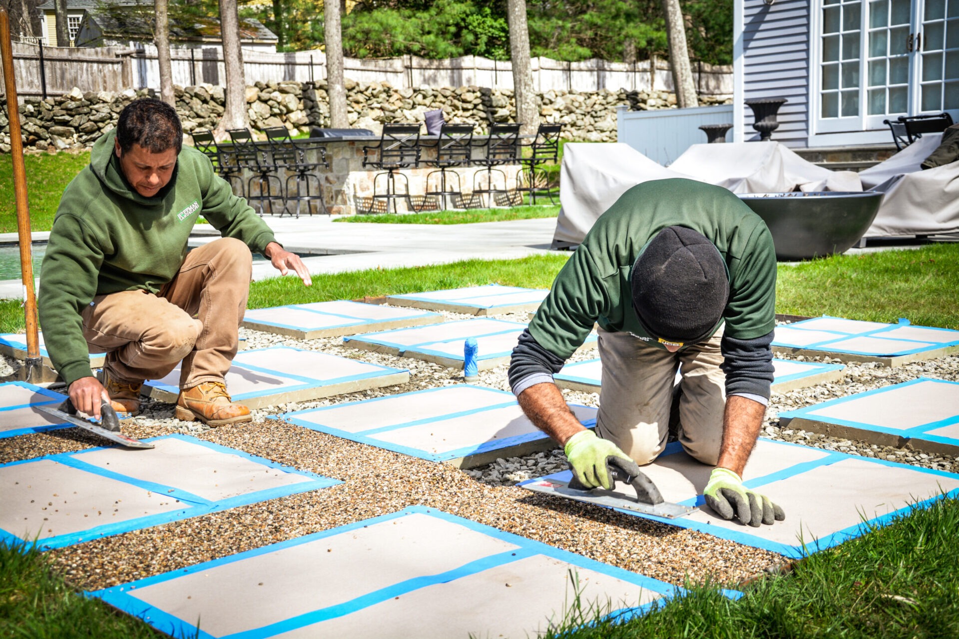 Two people are installing square pavers in a backyard, surrounded by greenery and outdoor furniture, on a sunny day with clear skies.