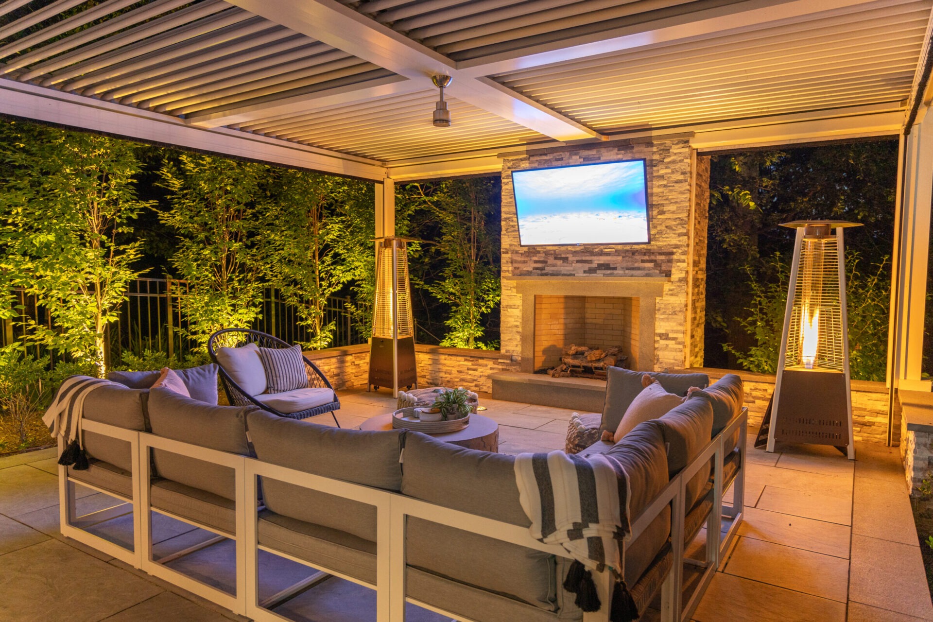 An outdoor living space at dusk with comfortable seating, a fireplace, television, heating lamps, and lush greenery visible beyond the pergola roof.