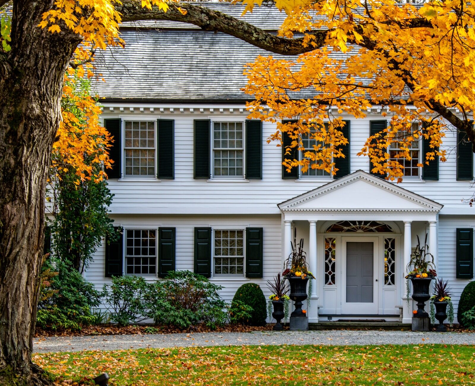 A classic two-story white house with black shutters, autumn leaves, a portico entrance, and decorative planters under a bright fall canopy.