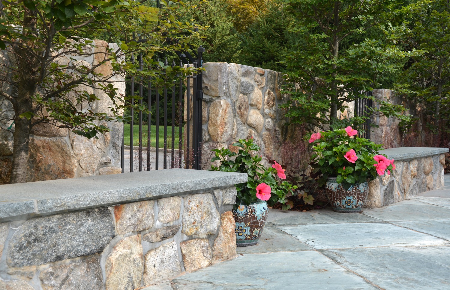 A stone wall with integrated benches borders a walkway, adorned with potted pink flowers, behind a metal gate, surrounded by greenery and trees.