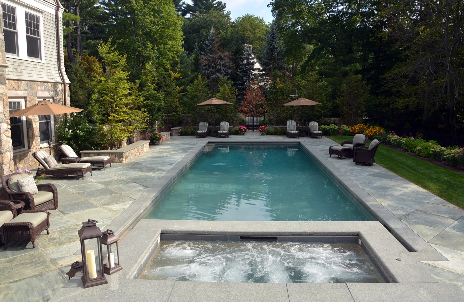 An elegant outdoor pool with an attached hot tub, flanked by lounge chairs and umbrellas, set against a backdrop of lush greenery and a stone house.
