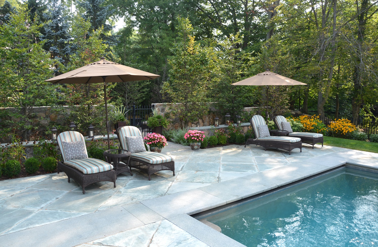 An outdoor pool area with lounge chairs, umbrellas, and flowering plants surrounded by trees with a stone wall in the background.