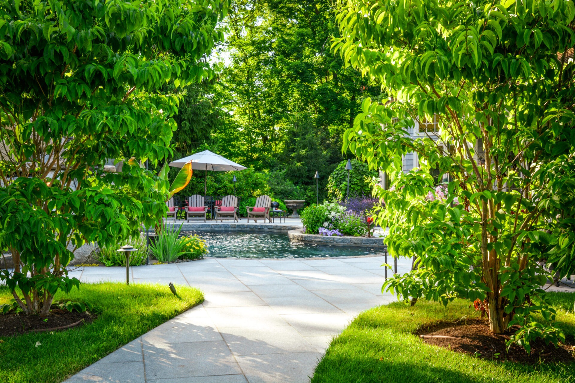 A tranquil garden setting featuring a swimming pool, lounge chairs under an umbrella, lush greenery, landscaped plants, and a paved walkway bathed in sunlight.