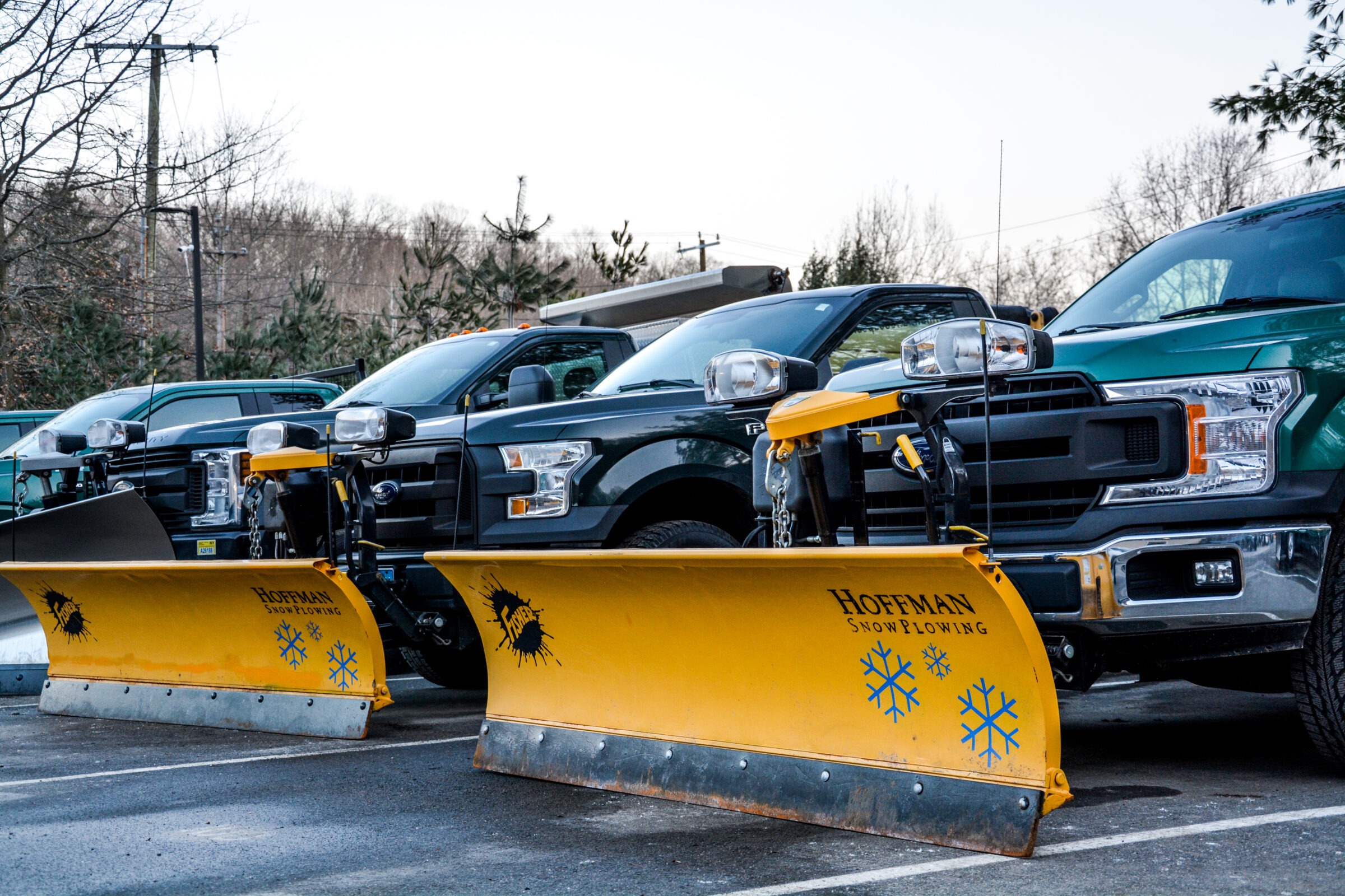 A line of pickup trucks equipped with yellow snowplow attachments, branded "Hoffman Snowplowing," parked in a lot with bare trees in the background.