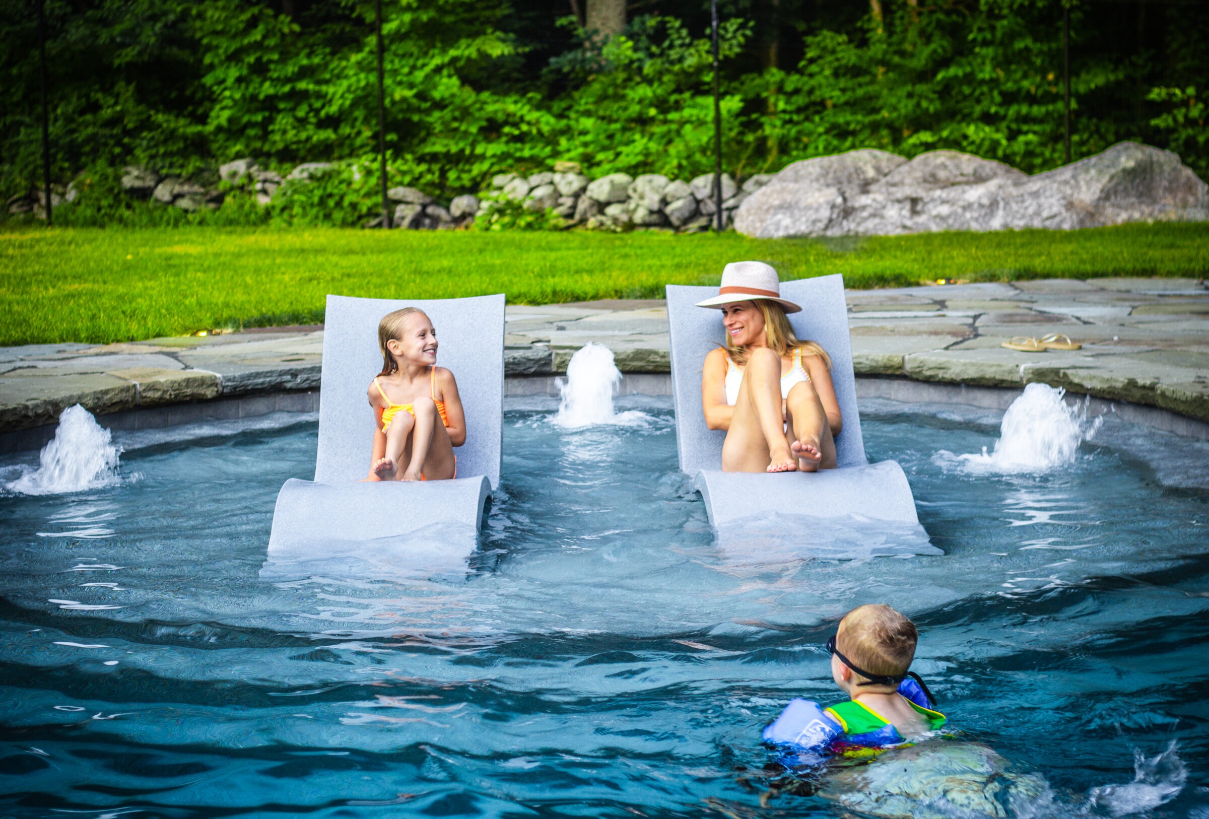 Two people relax on floating chairs in a pool, with a child nearby. They enjoy a sunny day surrounded by natural greenery and water features.