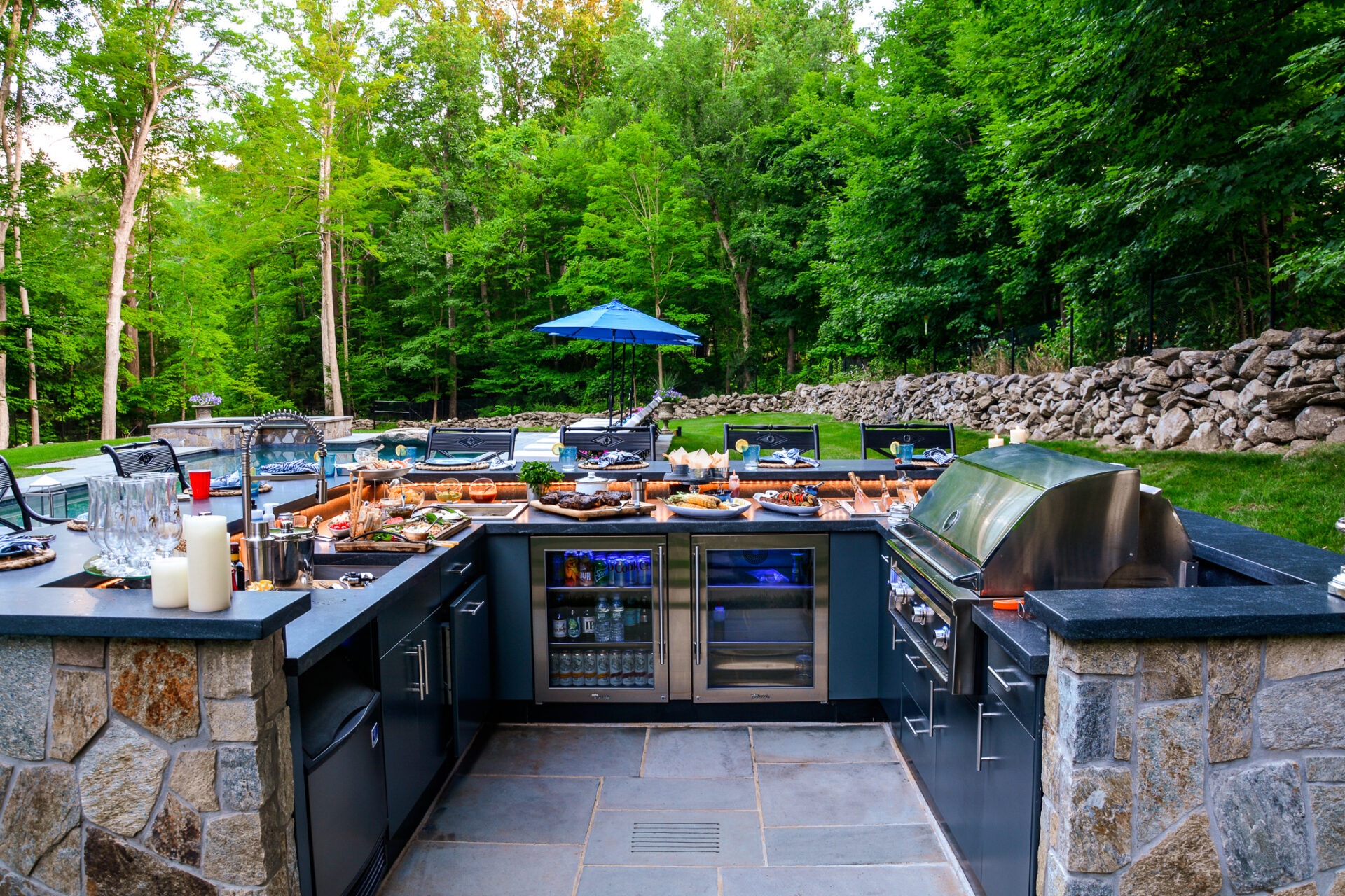 An outdoor kitchen with barbecue grills and a variety of foods set in a patio surrounded by lush greenery and a stone wall.