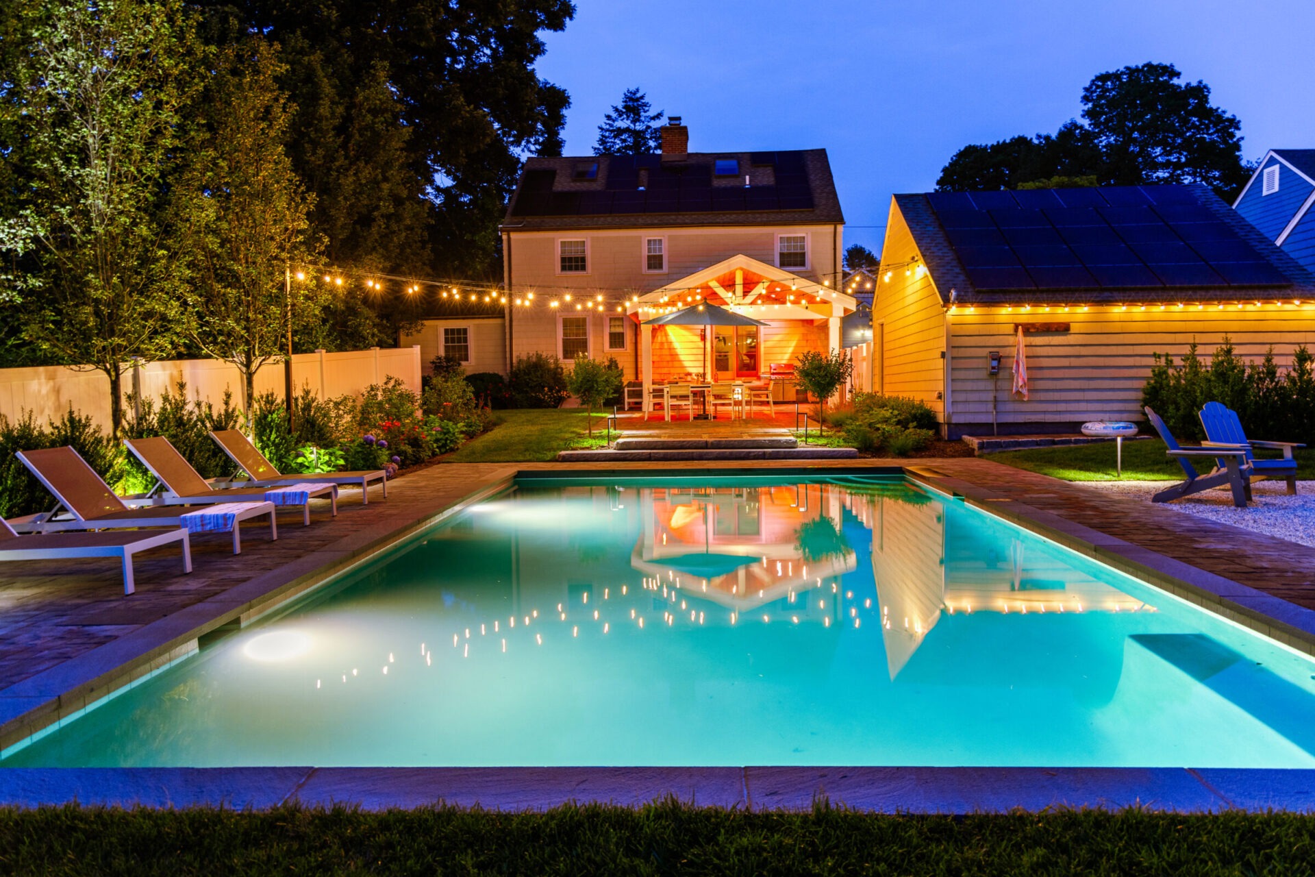 A tranquil evening setting featuring an illuminated swimming pool, string lights, loungers, and a house with a patio canopy. The environment is serene and inviting.