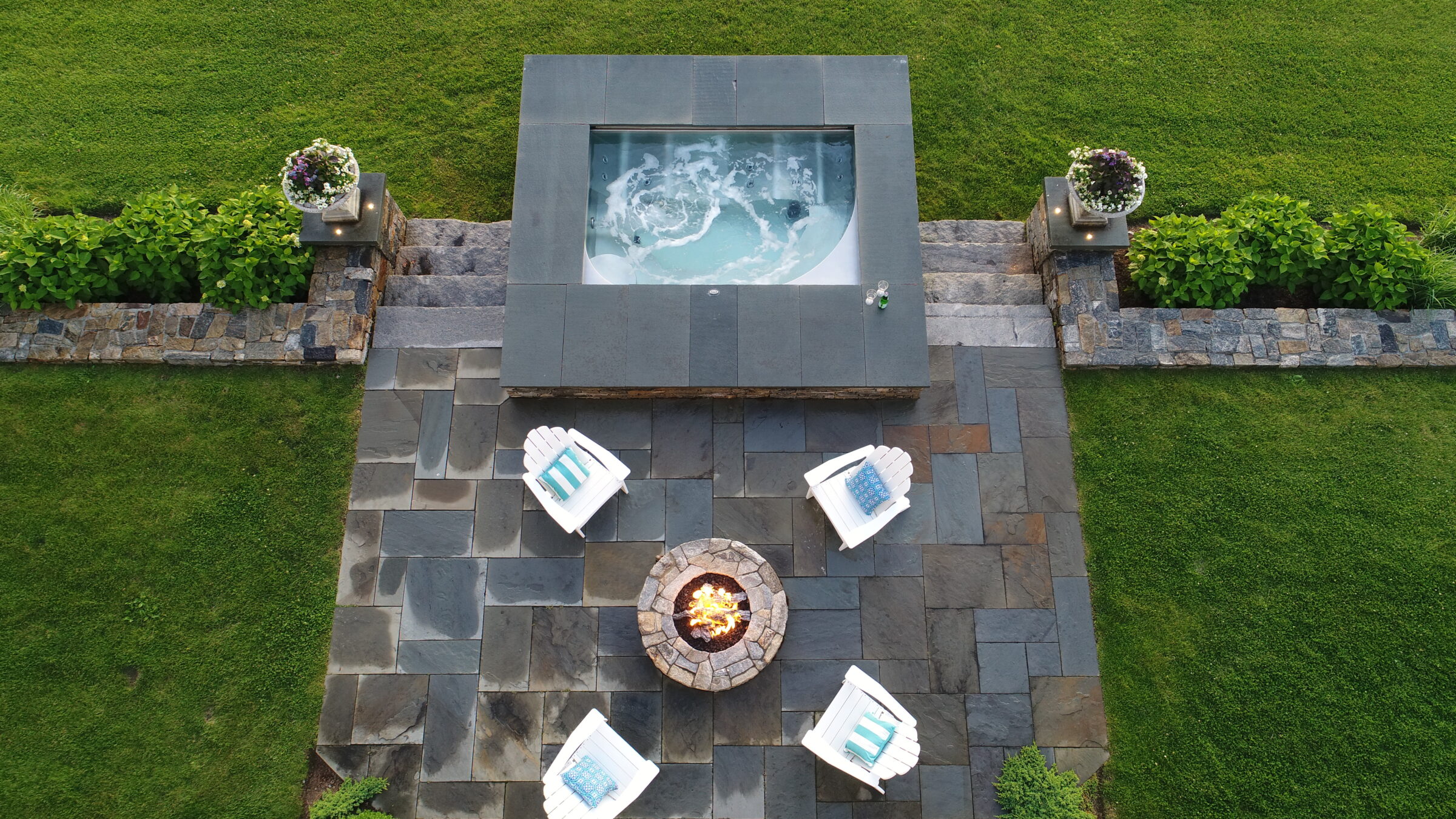 An aerial view of a backyard with a jacuzzi, a fire pit, and four Adirondack chairs, surrounded by well-maintained grass and stone walls.
