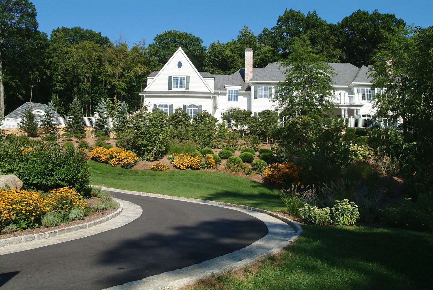 A large white house with a landscaped garden, paved driveway, lush greenery, and clear blue skies on a sunny day. No people are visible.
