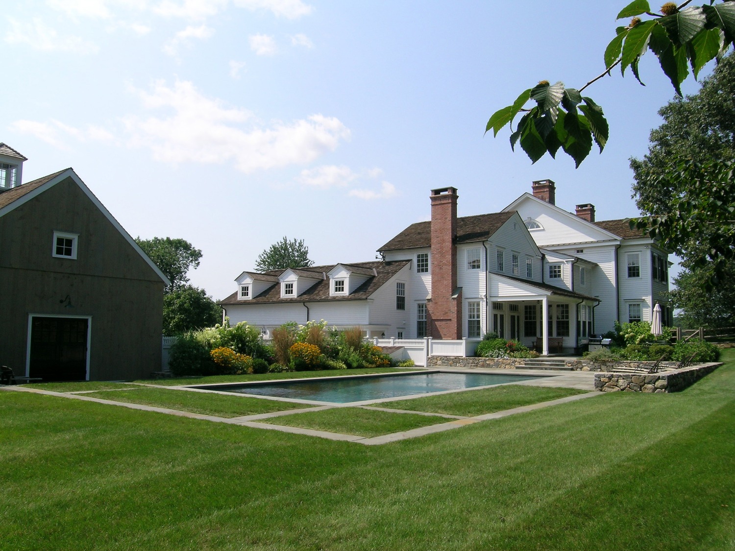 This image shows a large white house with multiple chimneys, a swimming pool, a green lawn, and an adjacent grey barn-like structure under a clear sky.
