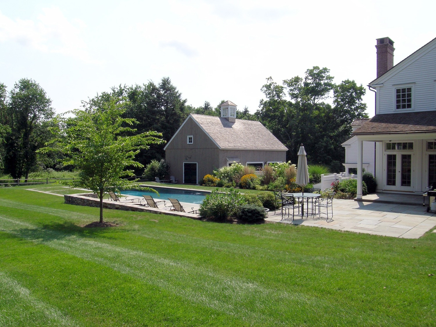 This image shows a lush backyard with a swimming pool, a patio area with furniture, a well-manicured lawn, and traditional-style houses under a clear sky.
