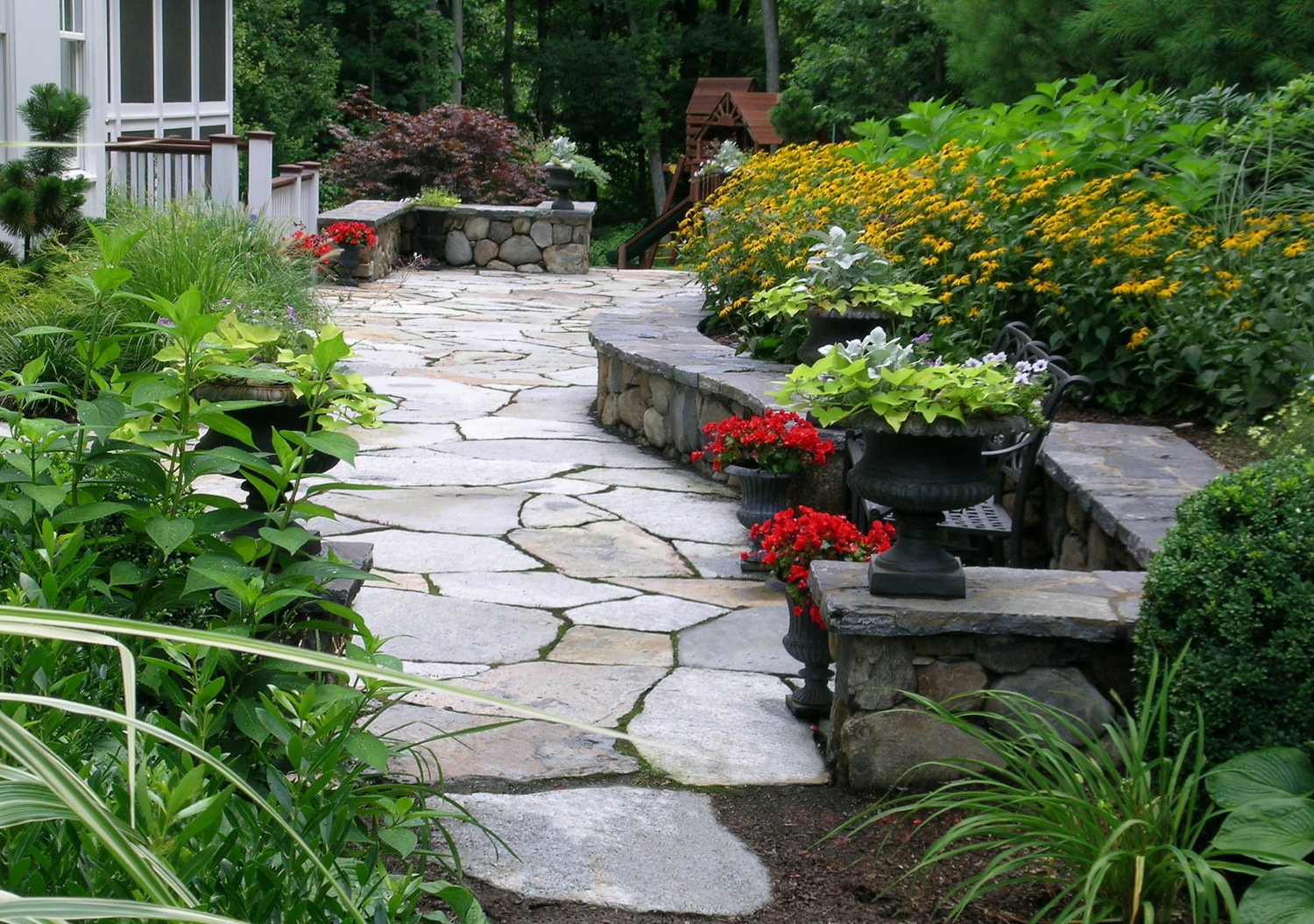 A beautiful garden pathway lined with vibrant flowers and lush greenery, featuring stone pavers and decorative planters, leads to a charming white house.