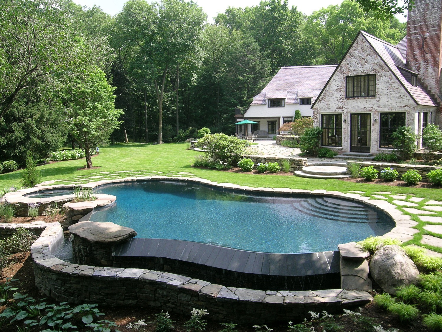 An elegantly landscaped backyard with a curvy swimming pool, stone borders, manicured lawns, and a charming house with large windows and a sloping roof.
