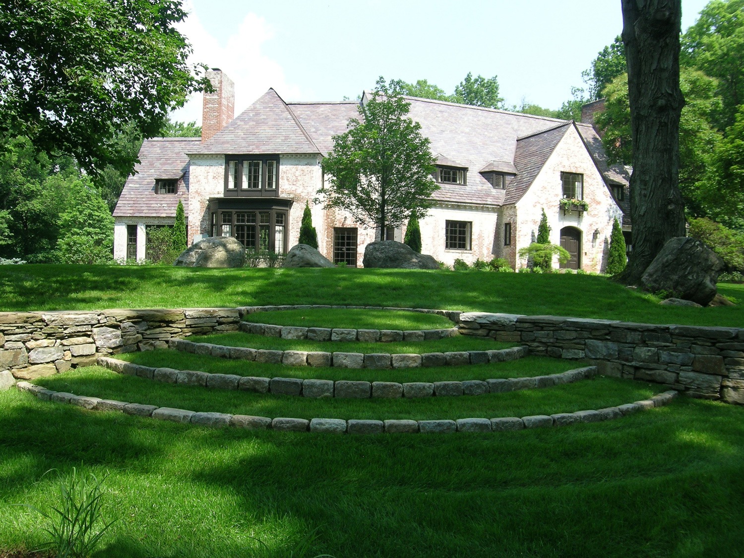 A large stone house with a slate roof, surrounded by lush greenery, featuring stone steps leading up to a well-manicured lawn and trees.