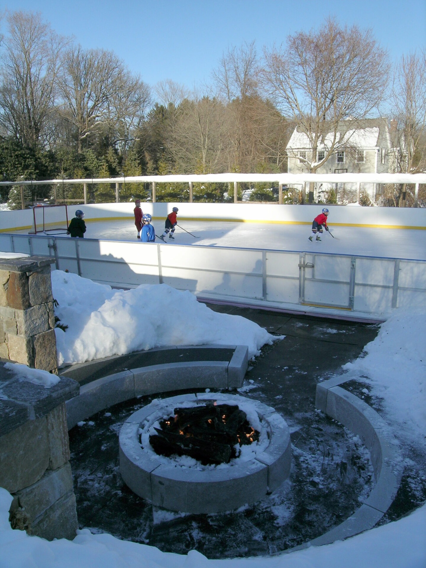 An outdoor ice rink surrounded by snow with four children skating, protected by helmets. A lit fire pit is visible in the foreground, with trees and a house in the background.