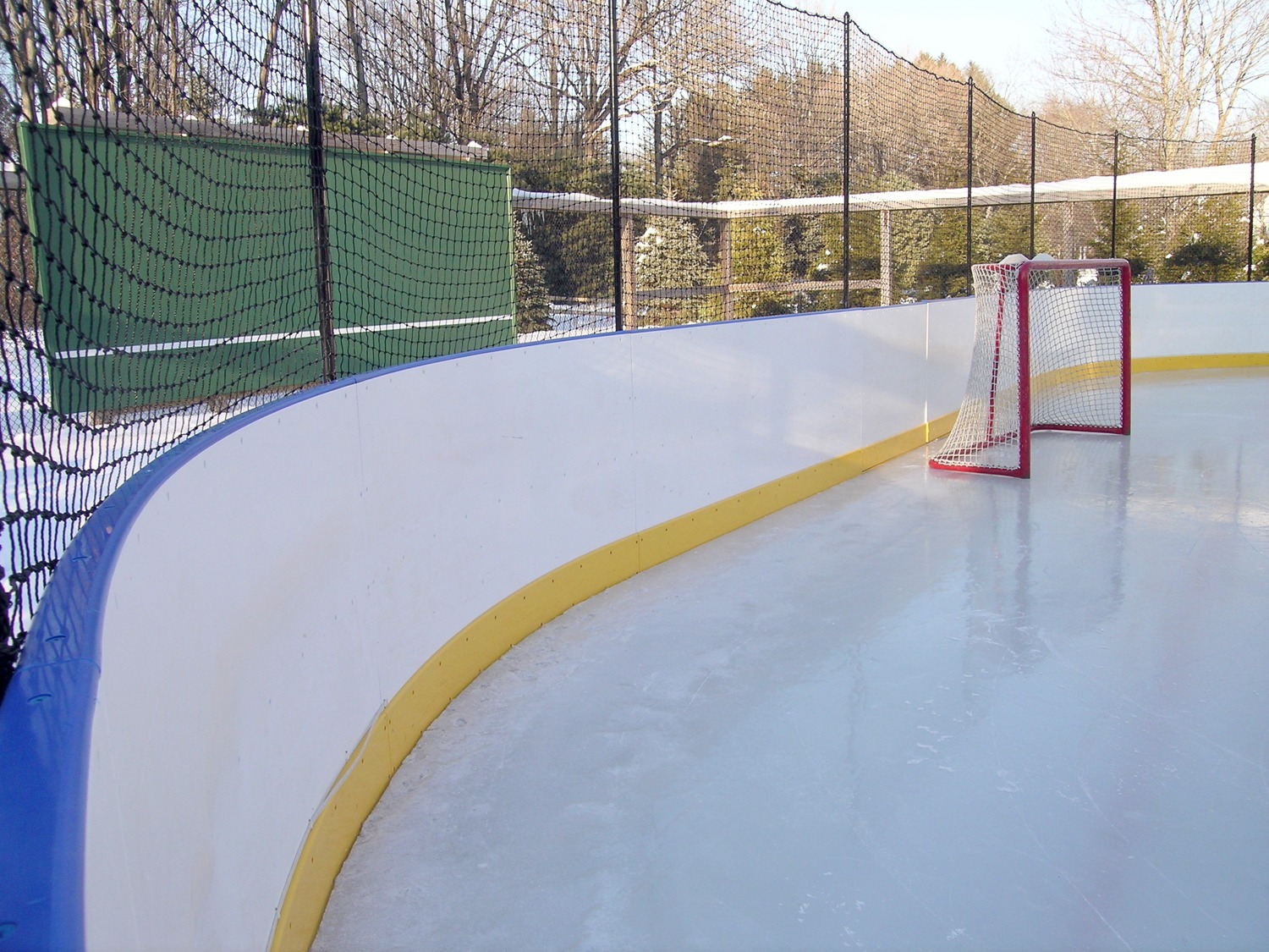 Empty outdoor hockey rink with a red goalpost, white ice surface, yellow kick plate, and netting against a backdrop of trees under a clear sky.