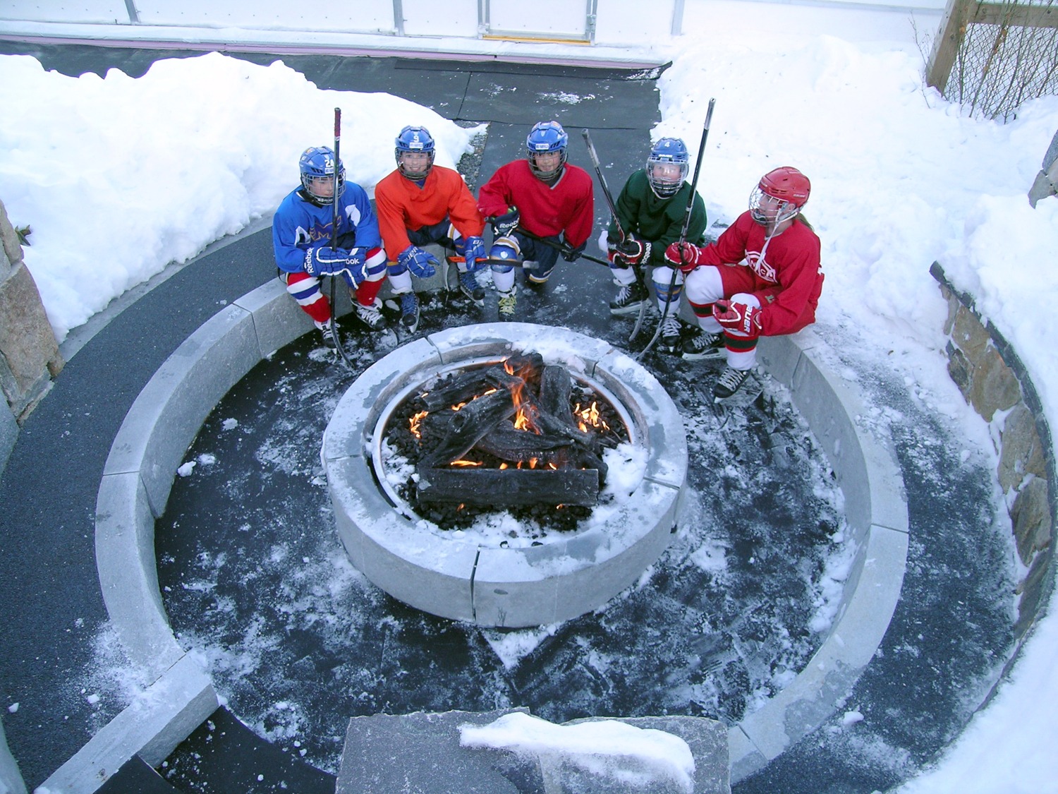 Five people in hockey gear sit around an outdoor fire pit surrounded by snow, resting their hockey sticks on the ground, in a wintry setting.