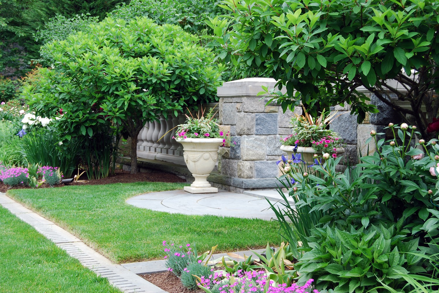 A well-manicured garden with lush greenery, flowering plants, a classic urn planter, stone pathways, and an elegant stone bench.