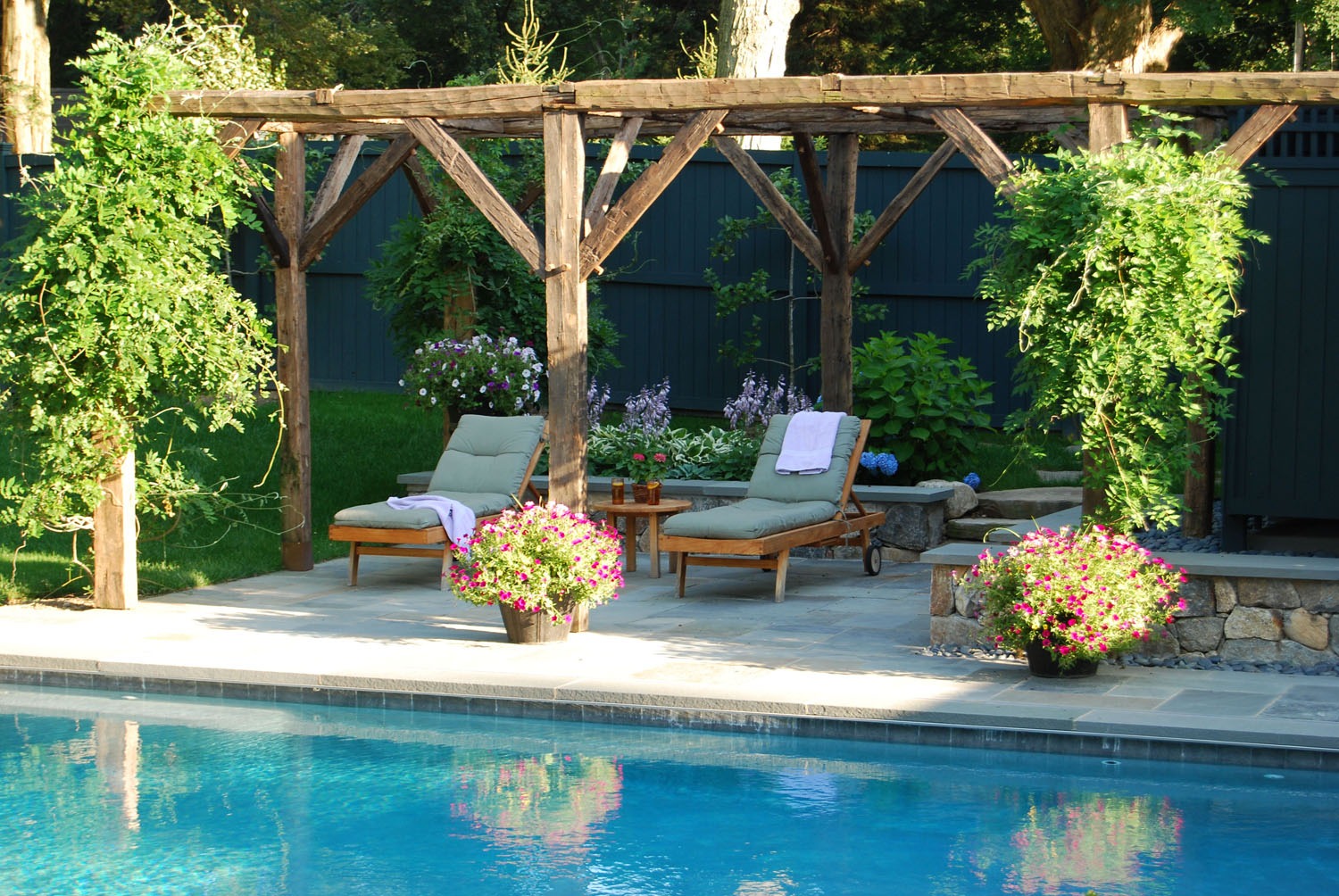 A serene backyard with a swimming pool, wooden sun loungers, vibrant flowers, and a pergola. The setting conveys leisure and tranquility, surrounded by greenery.