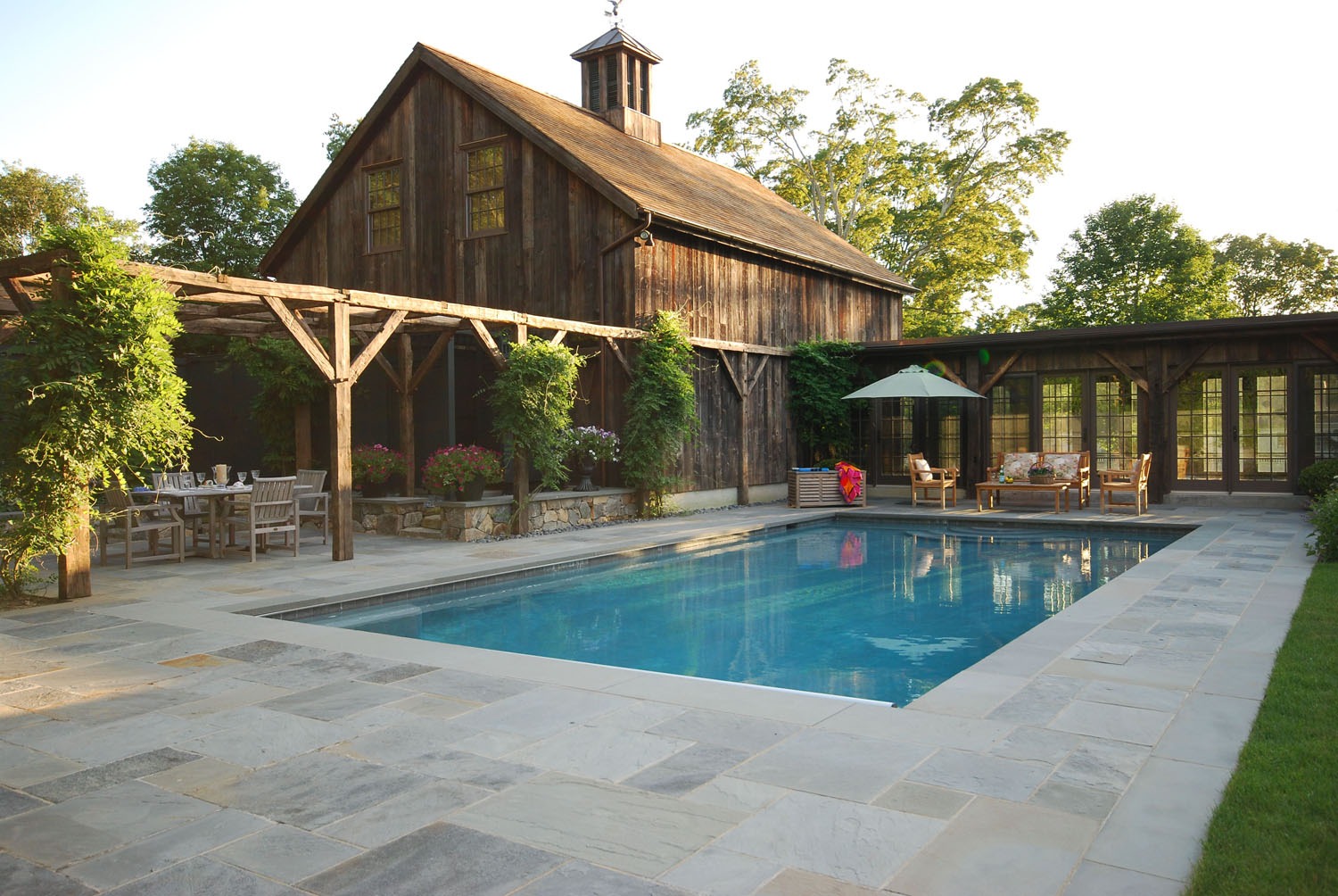 This image features a serene backyard with a swimming pool, rustic barn-style architecture, a pergola, outdoor furniture, and lush greenery on a sunny day.