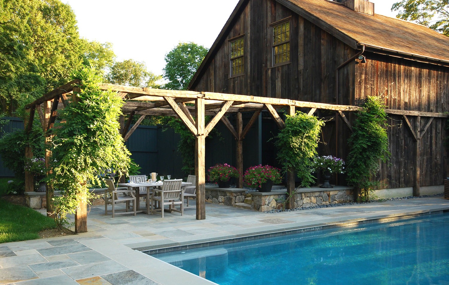 A rustic wooden barn with a pergola, creeper plants, an outdoor dining set, and a swimming pool in a lush garden setting at dusk.