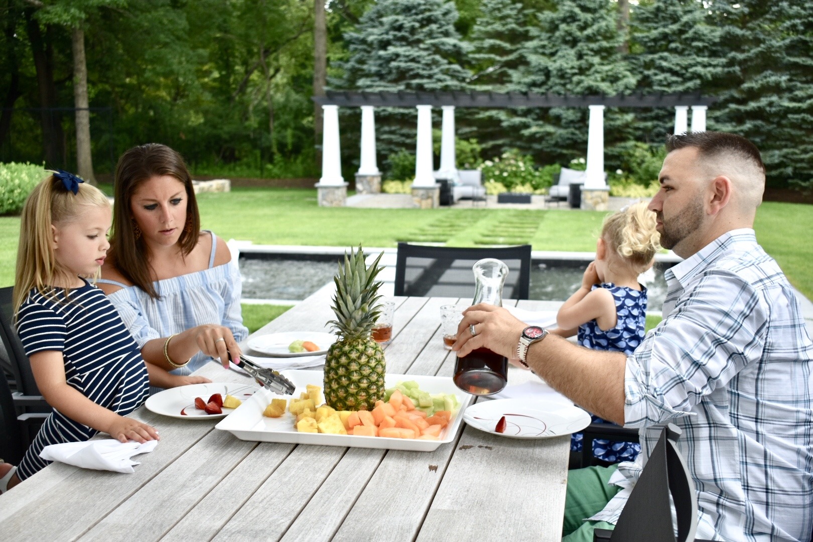 A family of four enjoys an outdoor meal together. A person is pouring a drink, while another adult helps a child with food.