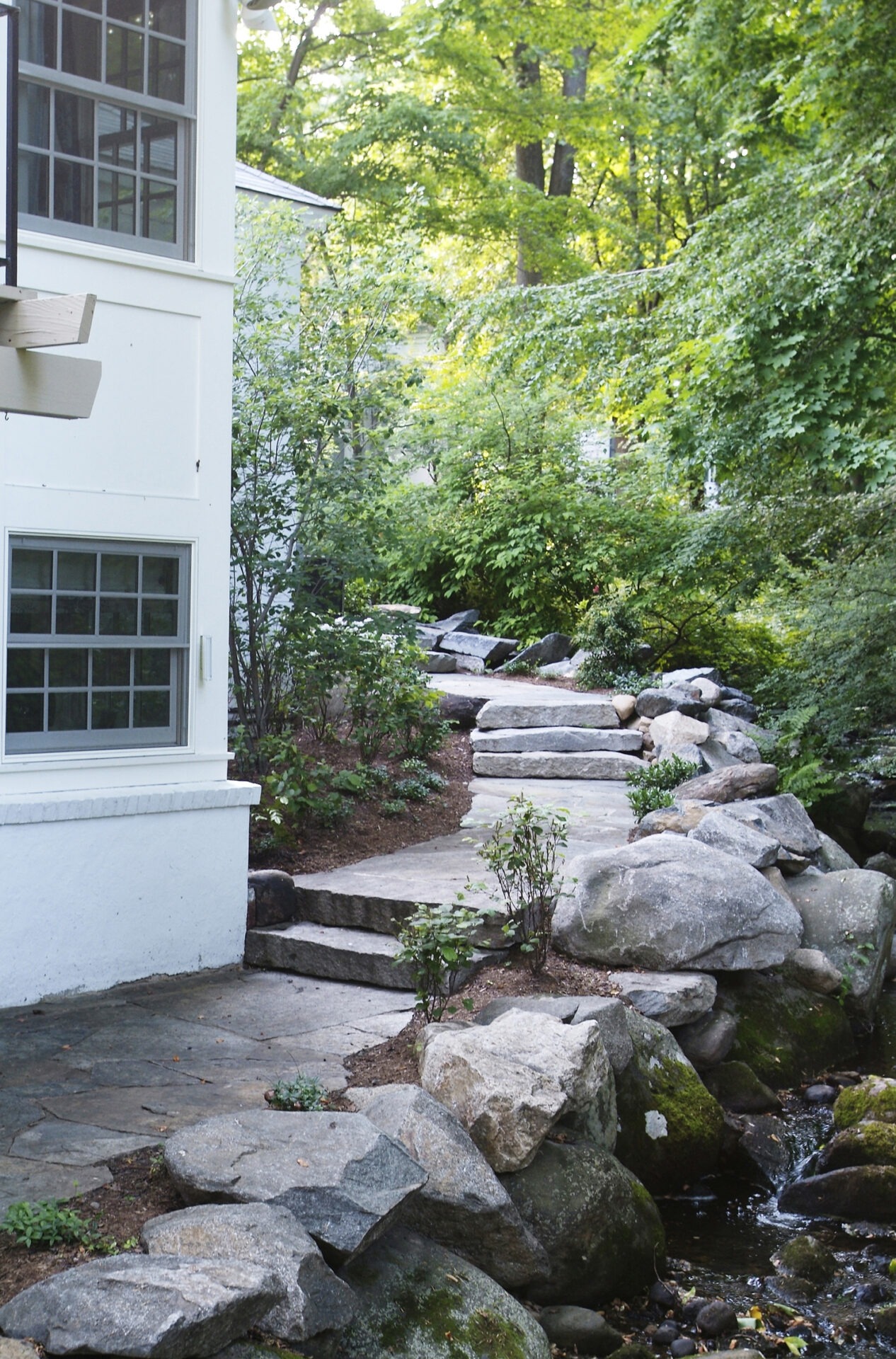 A serene garden path with large stepping stones leads past a small stream, adjacent to a white building with windows, amidst lush greenery.