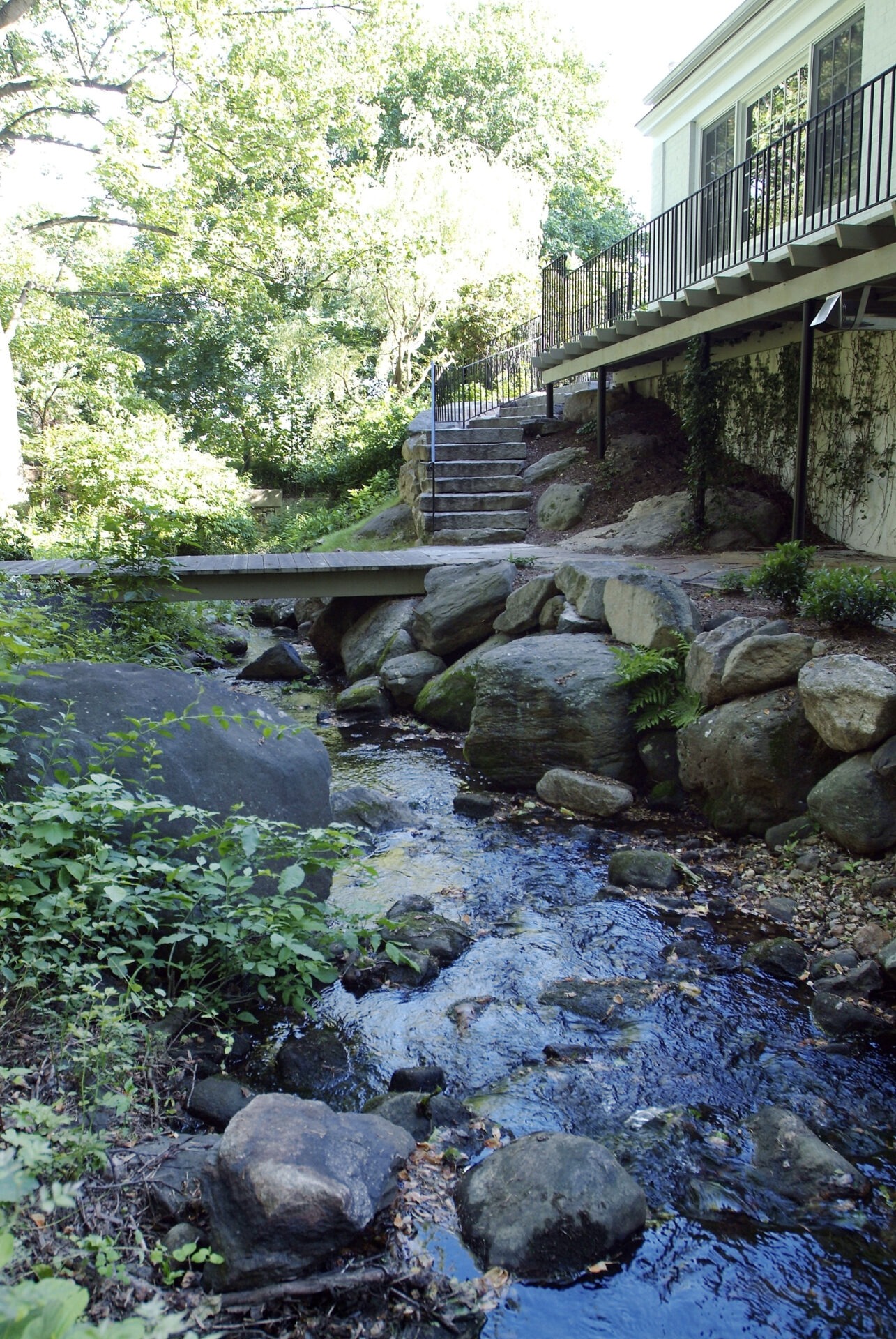 A serene stream flows between rocky banks beneath a wooden bridge, leading to stone steps by a building with a balcony amidst lush greenery.