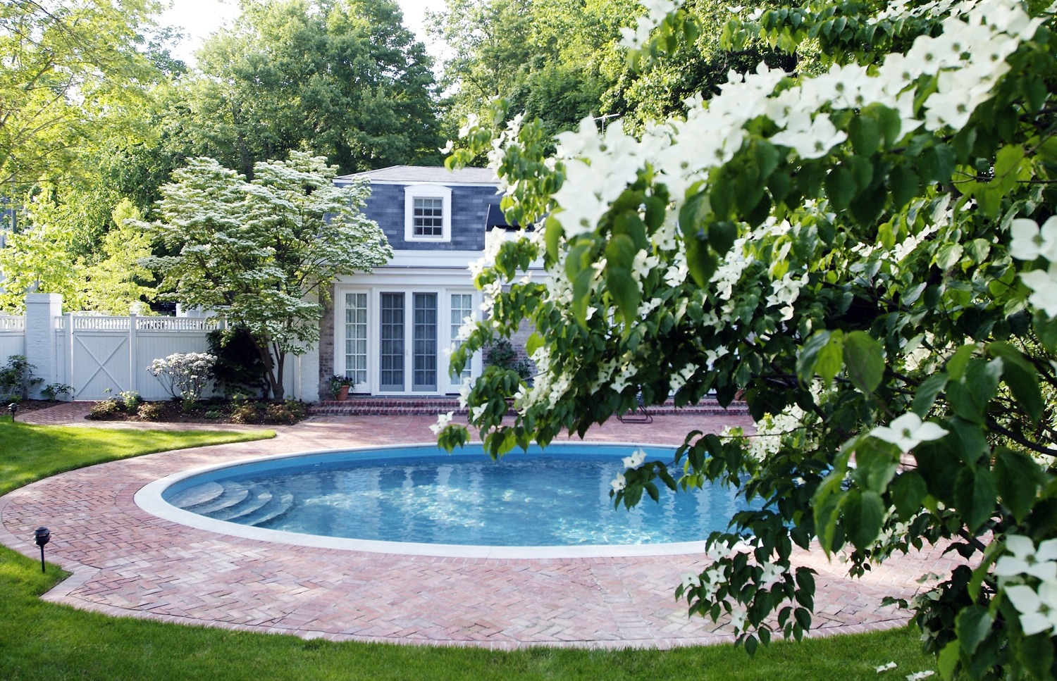Picture of a serene backyard with a kidney-shaped swimming pool, brick patio, dogwood flowers in the foreground, and a classic house with shutters.