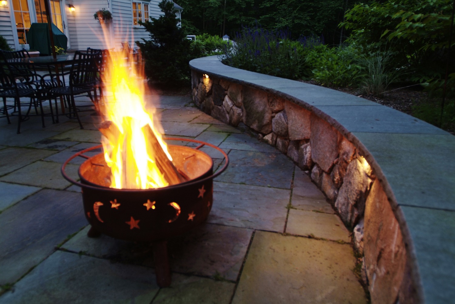 A metal fire pit with star cutouts, containing a blazing fire, placed on a stone patio near a stone curved wall, with a house in the background.