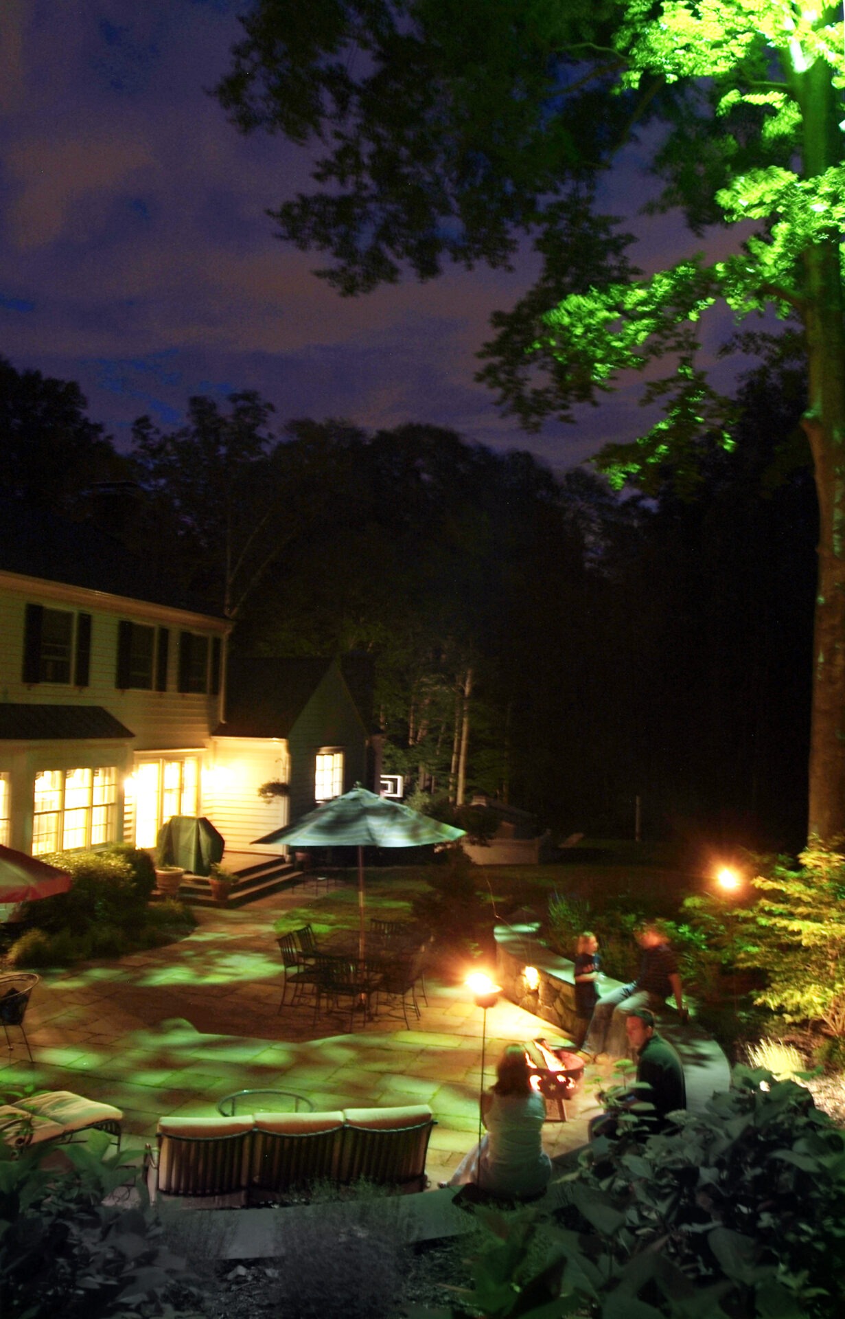 A nighttime scene in a backyard with illuminated house windows, patio furniture, a pool, and a group of people enjoying a fire pit.