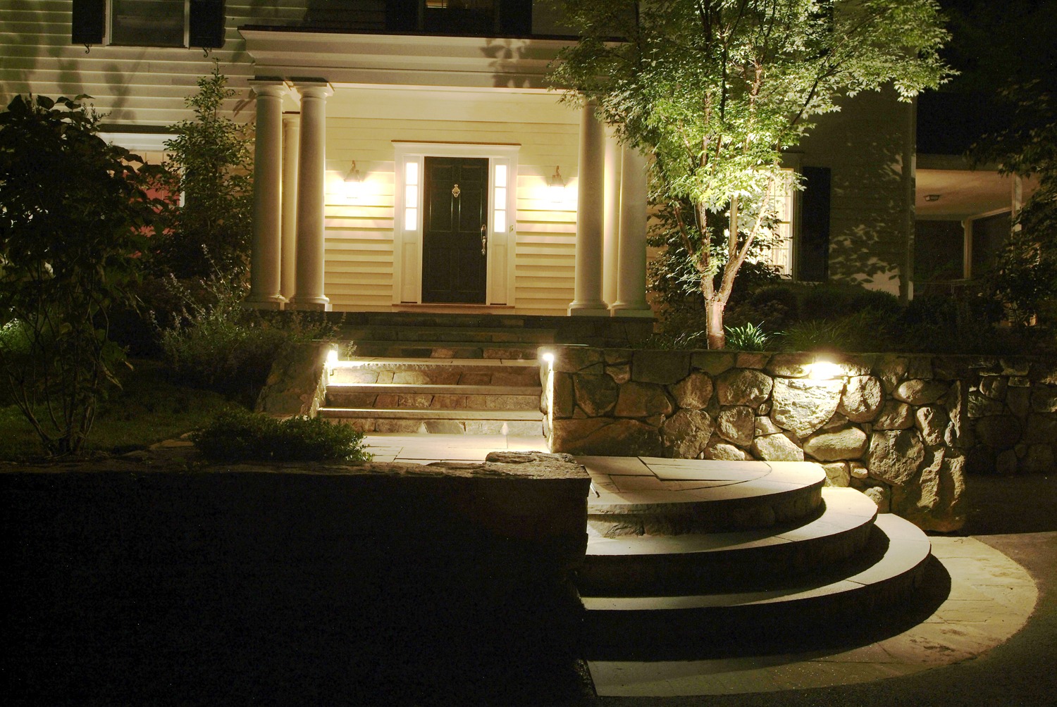 This image shows a house entryway at night with illuminated steps, classical pillars, a front door, landscaping, and exterior lights.