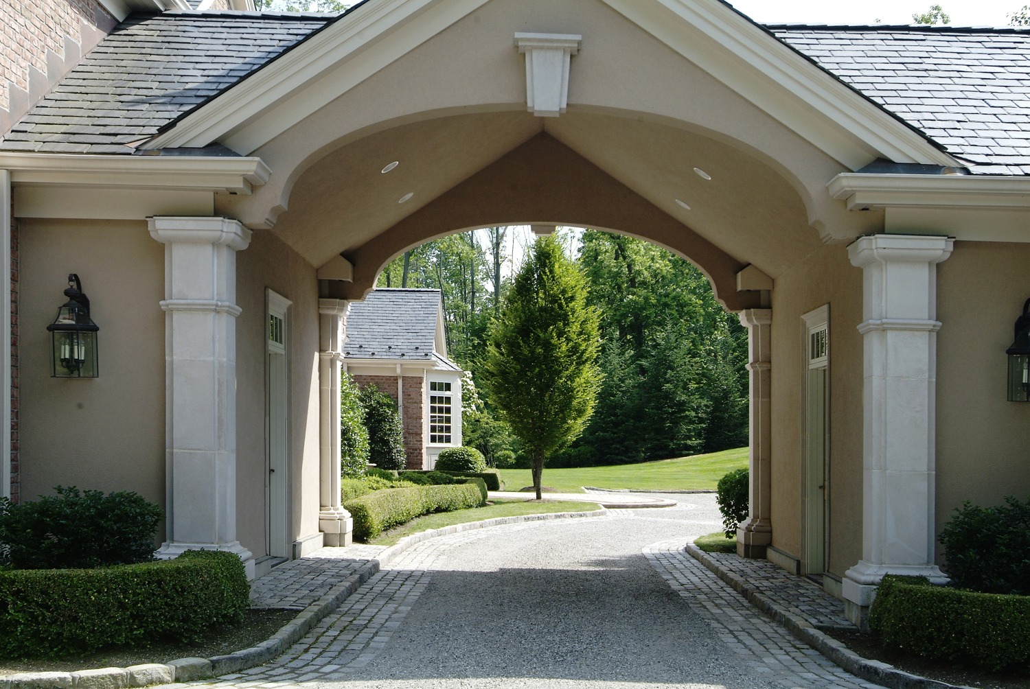 An elegant stone driveway passes through an arched porte-cochère, leading towards a manicured garden and a traditional brick building with greenery.