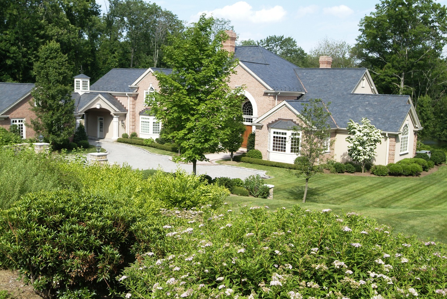 An expansive brick house with several chimneys and gables, lush landscaping, a manicured lawn, and a winding driveway in a sunny setting.