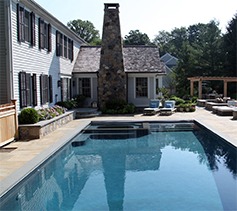 This image shows an outdoor swimming pool next to a traditional two-story house with white siding, accented by a stone chimney and patio area.