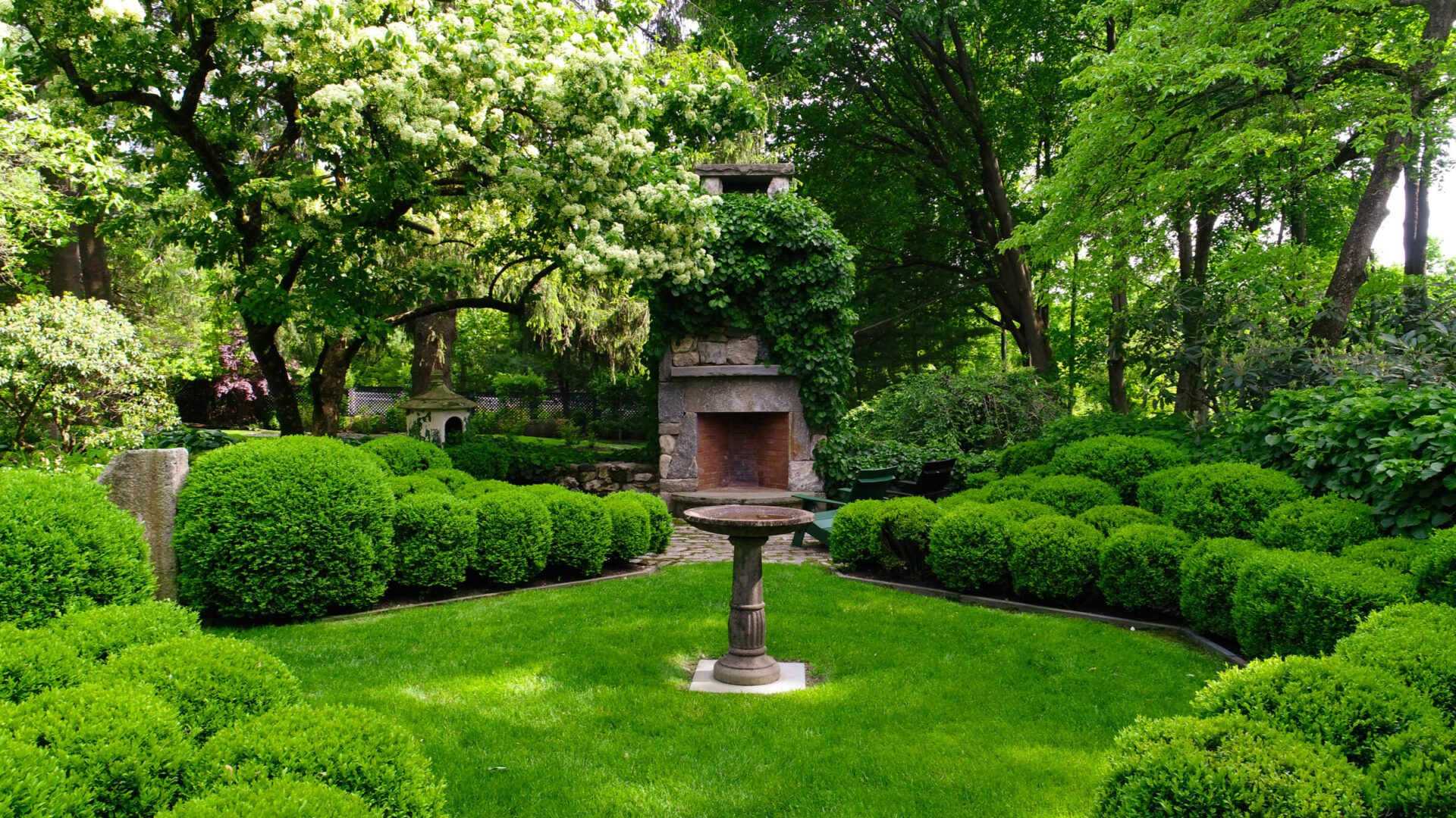 A lush garden with neatly trimmed bushes, a birdbath centerpiece, a stone fireplace covered in vines, surrounded by a variety of green trees.