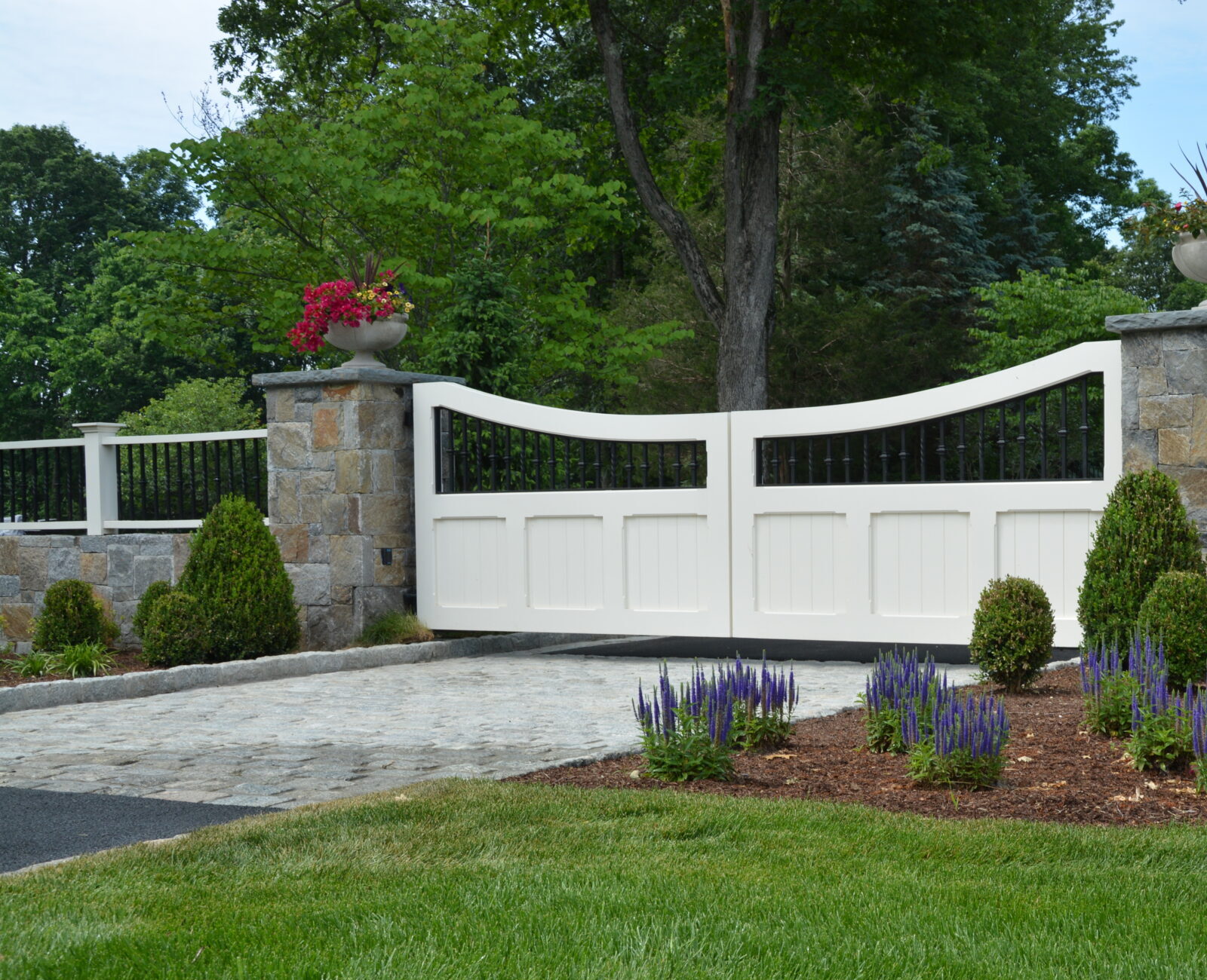 A white automated driveway gate with stone pillars and decorative plants stands between a lush lawn and a house partially visible in the background.