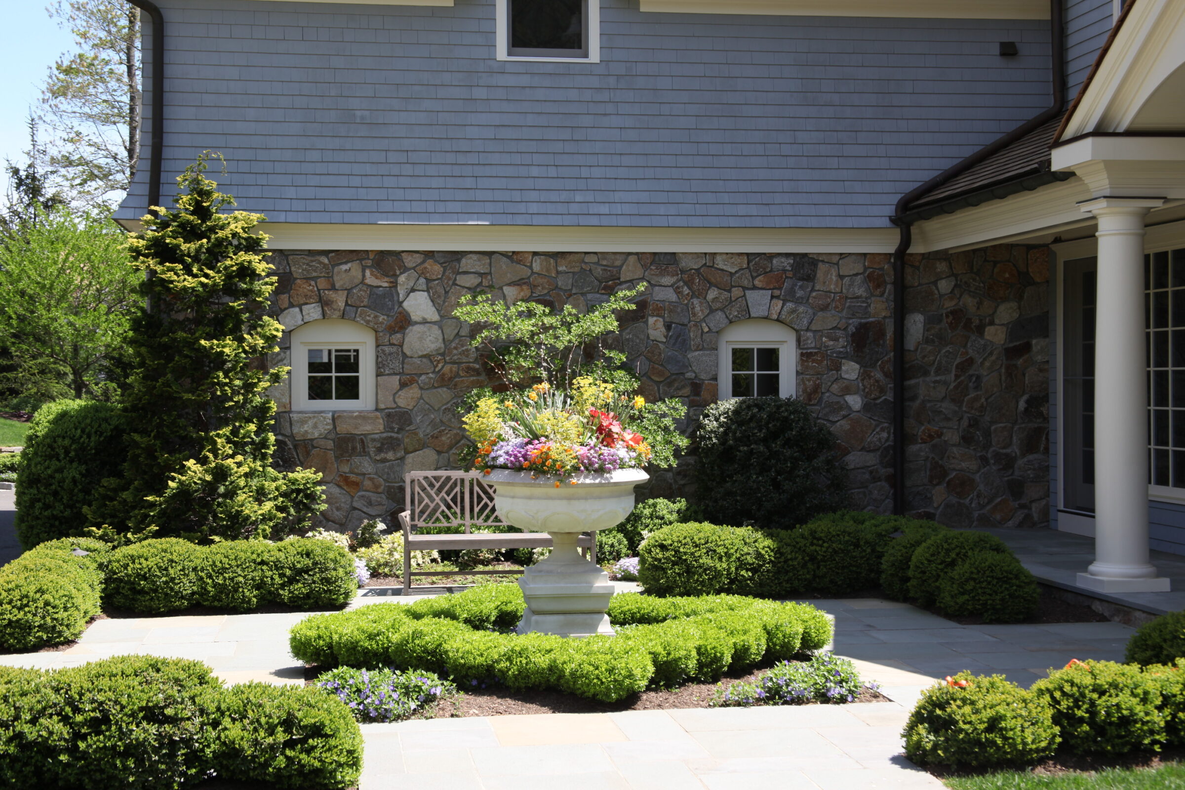 The image shows a landscaped garden in front of a house with stone facade, featuring a decorative planter with colorful flowers and trimmed bushes.