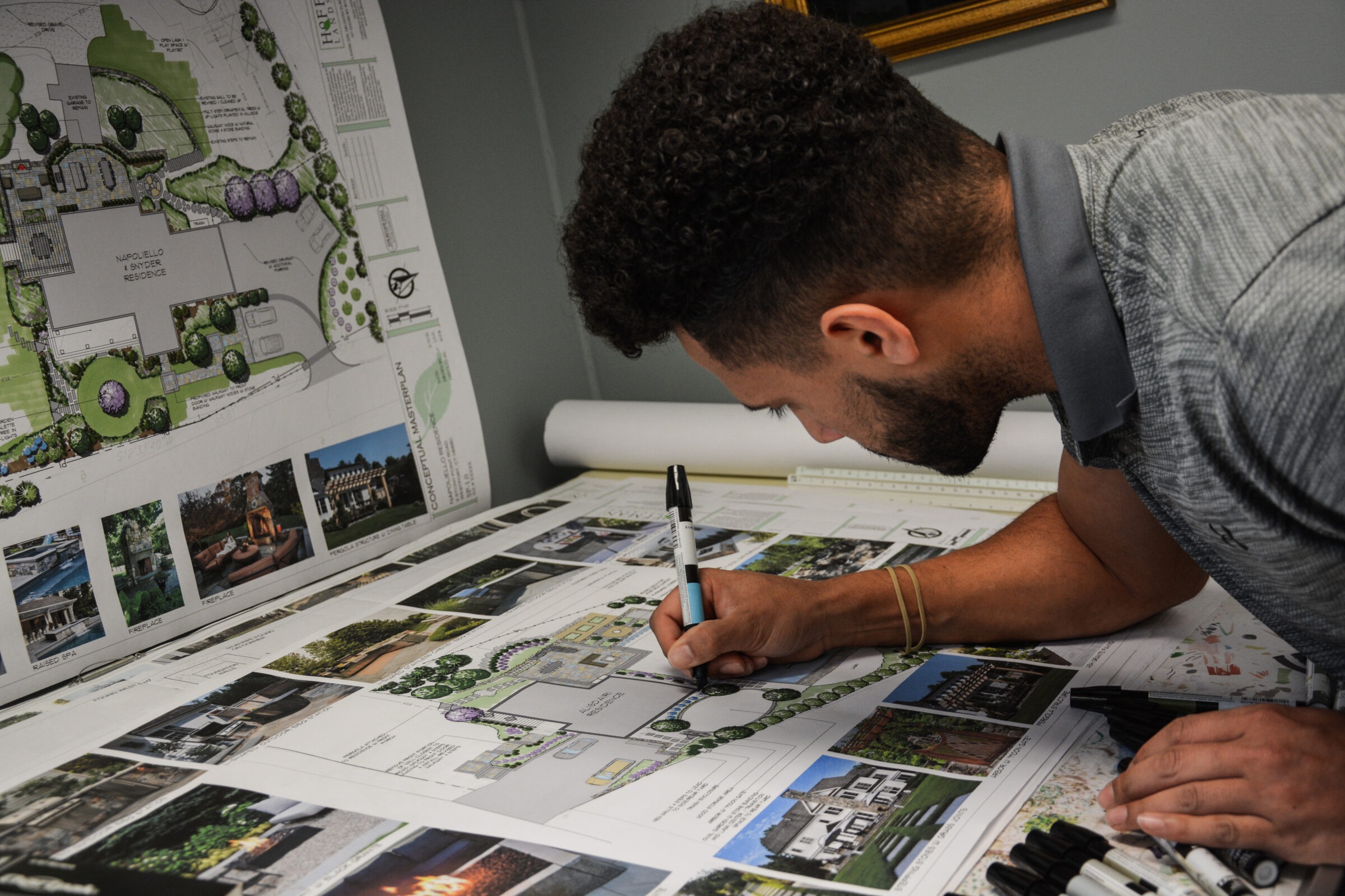 A focused person is drawing on a landscape design plan, surrounded by pens and printed images related to the project.