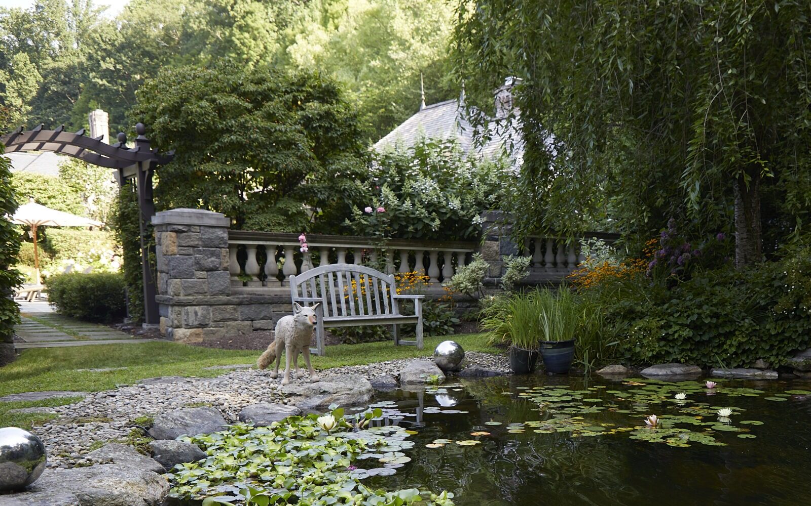 A tranquil garden with a pond full of water lilies, a waterfall, a dog standing on rocks, foliage around, and a garden bench in the background.