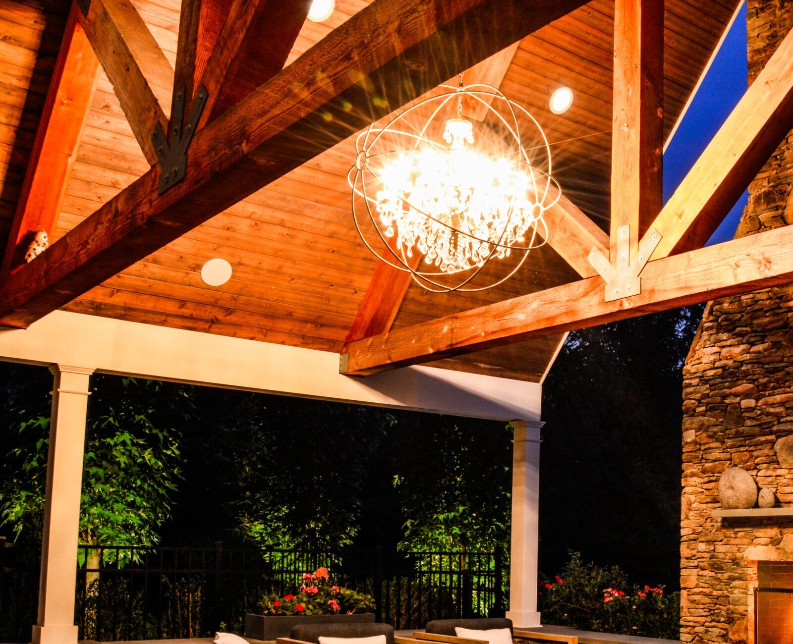 An elegant outdoor seating area featuring a stone fireplace, wooden beams, comfortable furniture, ambient lighting, and a view of a garden at dusk.
