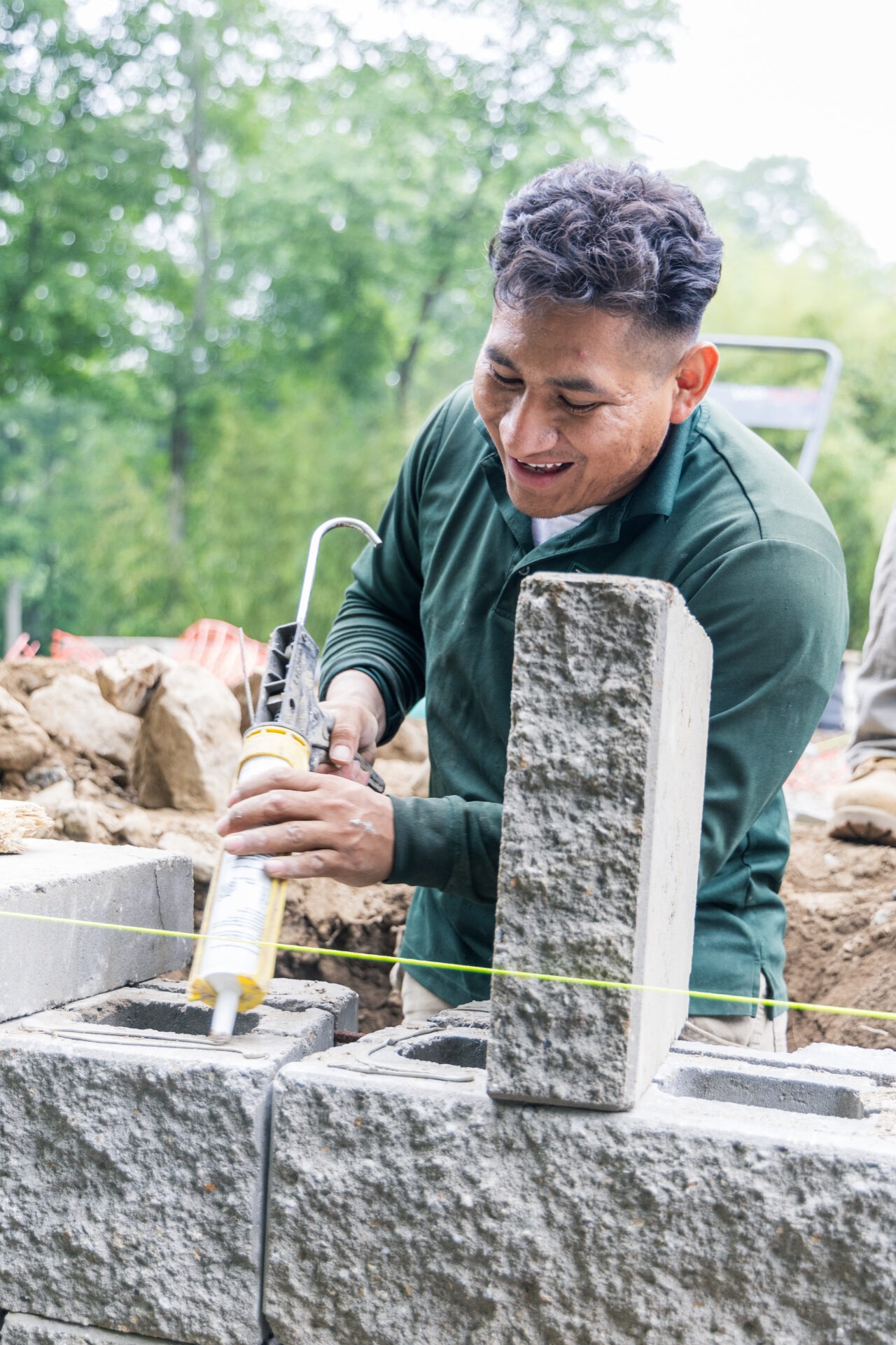 A person is applying adhesive from a caulk gun to concrete blocks, smiling while focused on construction work outdoors with greenery in the background.