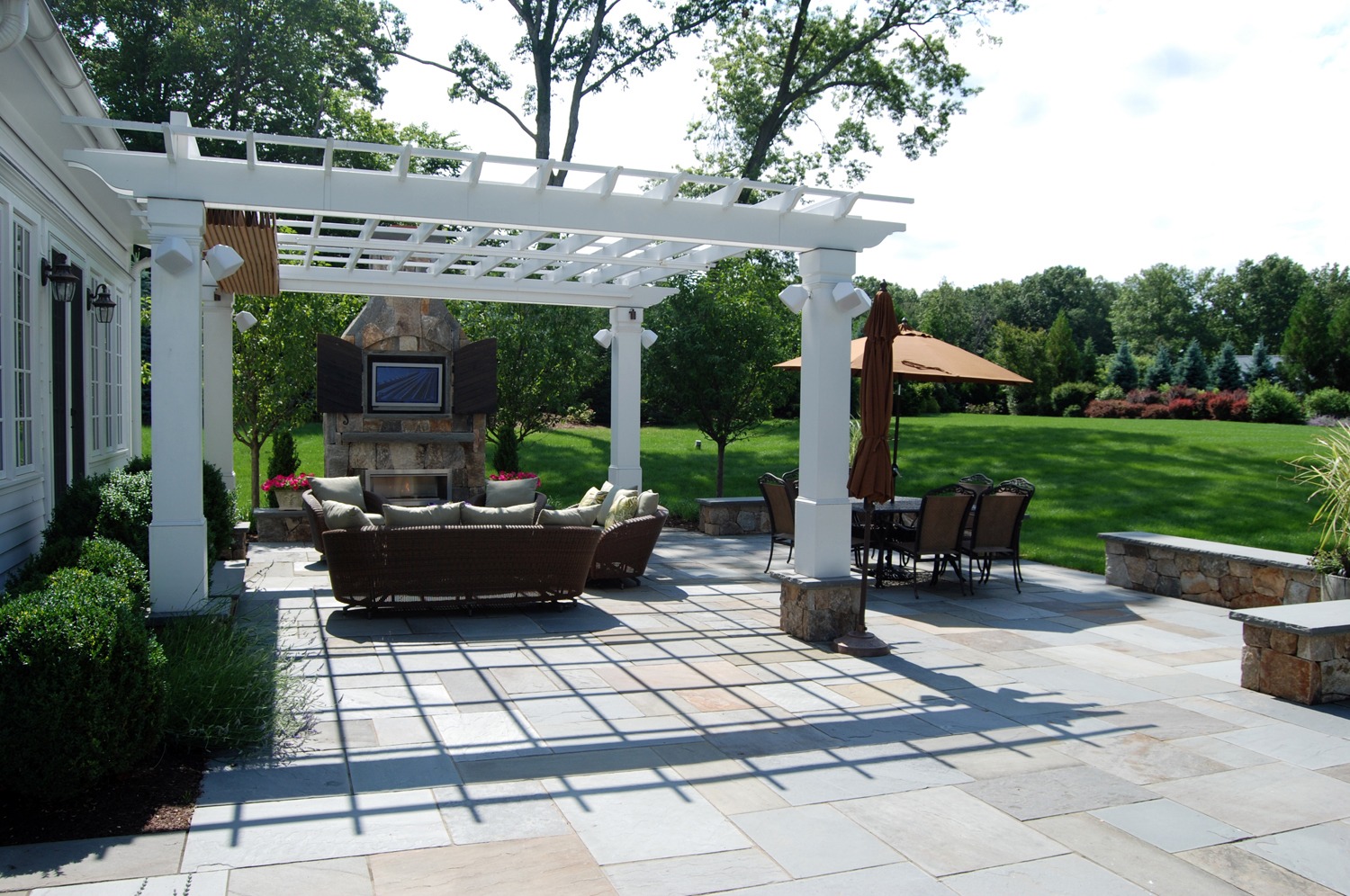 An elegant outdoor patio with a white pergola, stone fireplace, wicker sofa, dining set, and an umbrella, surrounded by lush greenery under a clear sky.