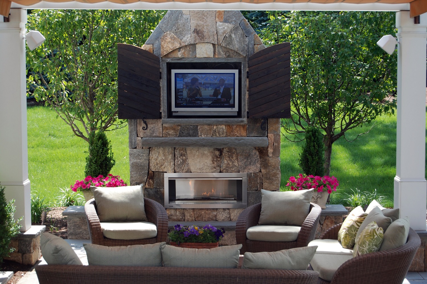 An outdoor sitting area with comfortable furniture, a stone fireplace, a mounted television, and vibrant plants, set against a verdant lawn backdrop.
