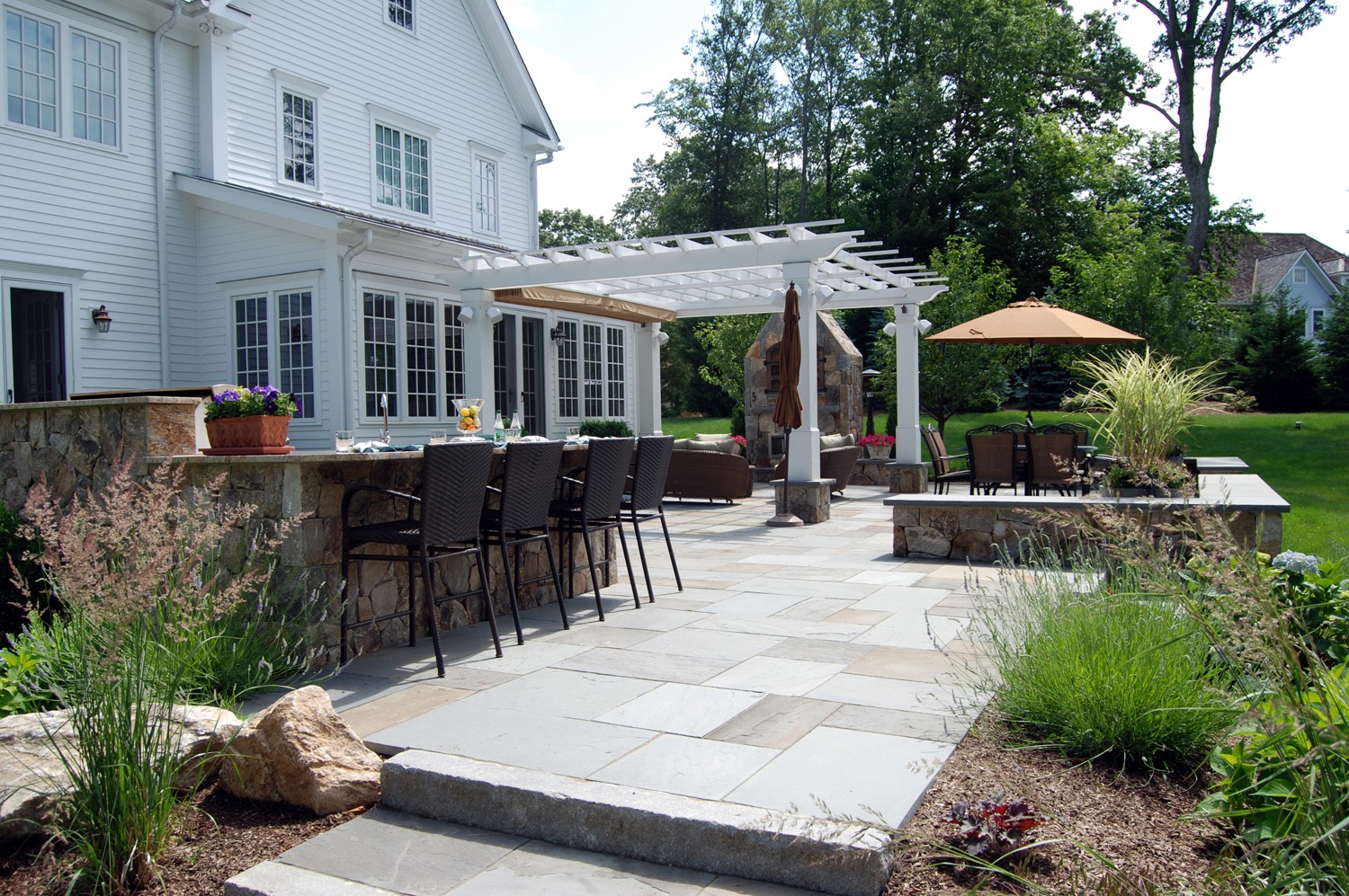 An elegant outdoor patio with a stone bar, seating area, pergola, and umbrella, adjacent to a white house with greenery and a clear sky.