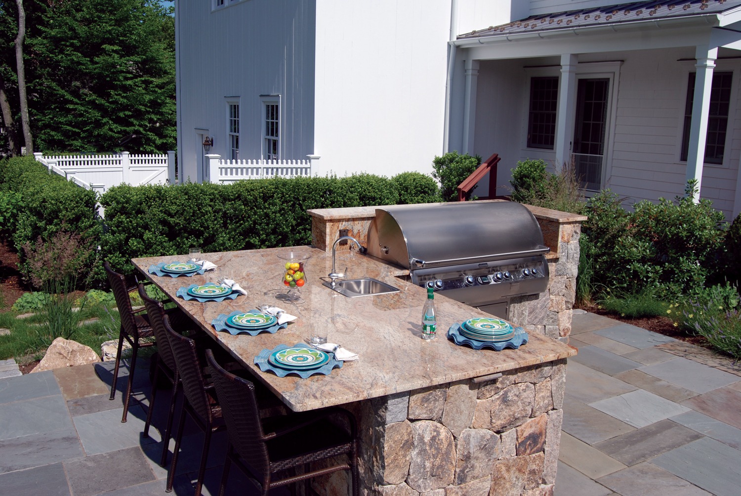 An outdoor kitchen with a built-in grill, sink, and stone countertop. Plates are set on the bar, and there's a white fence with greenery behind.